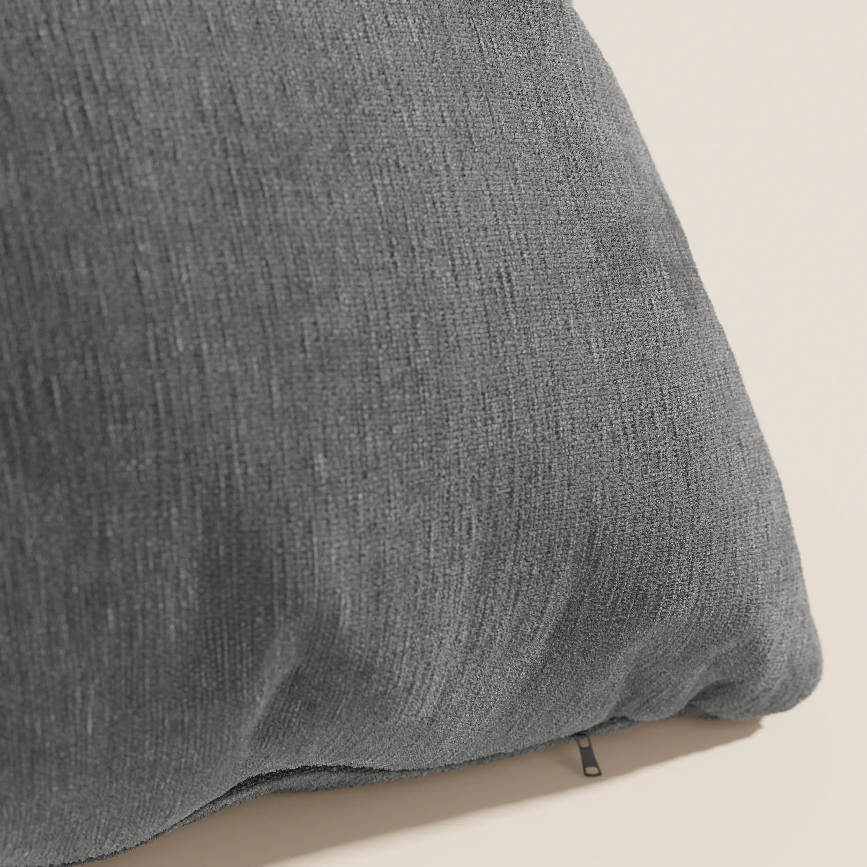 Un primo piano del cuscino Bolero di Divani.Store in morbido tessuto grigio scuro T31 antimacchia, impermeabile e non restringente con cerniera inferiore a vista, esposto su uno sfondo beige chiaro per un look accogliente e contemporaneo.