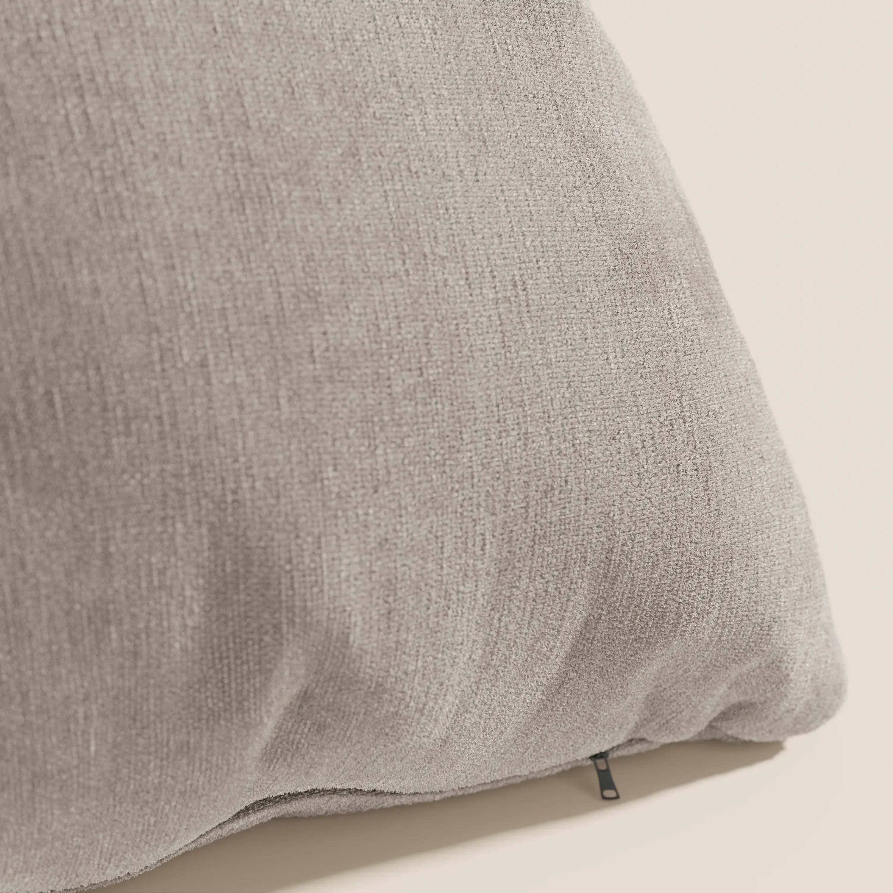Primo piano del cuscino Bolero di Divani.Store in grigio chiaro, con tessuto T31 morbido e testurizzato, resistente alle macchie, impermeabile e non restringente. Una piccola cerniera nera mostra il rivestimento sfoderabile su uno sfondo beige.