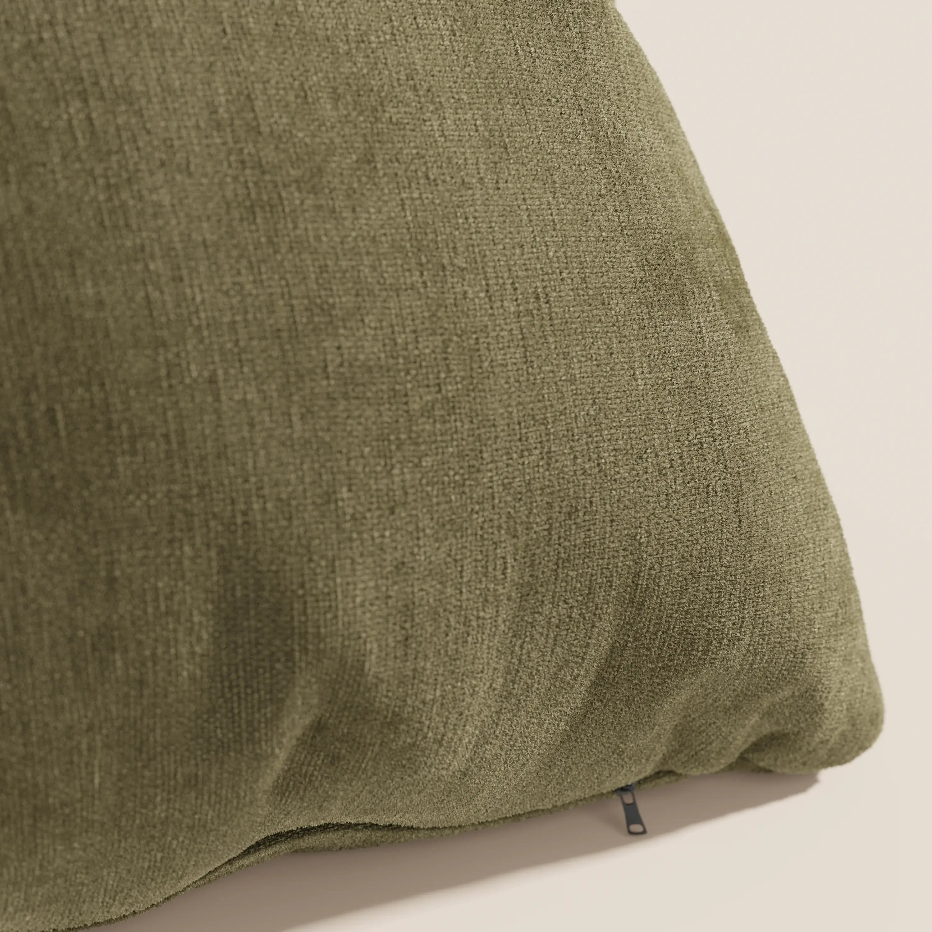 Primo piano del cuscino Bolero di Divani.Store in verde oliva, caratterizzato da un morbido tessuto T31 antimacchia, impermeabile e non restringente. In basso è visibile un piccolo tiretto nero per la chiusura lampo. Lo sfondo è beige chiaro.