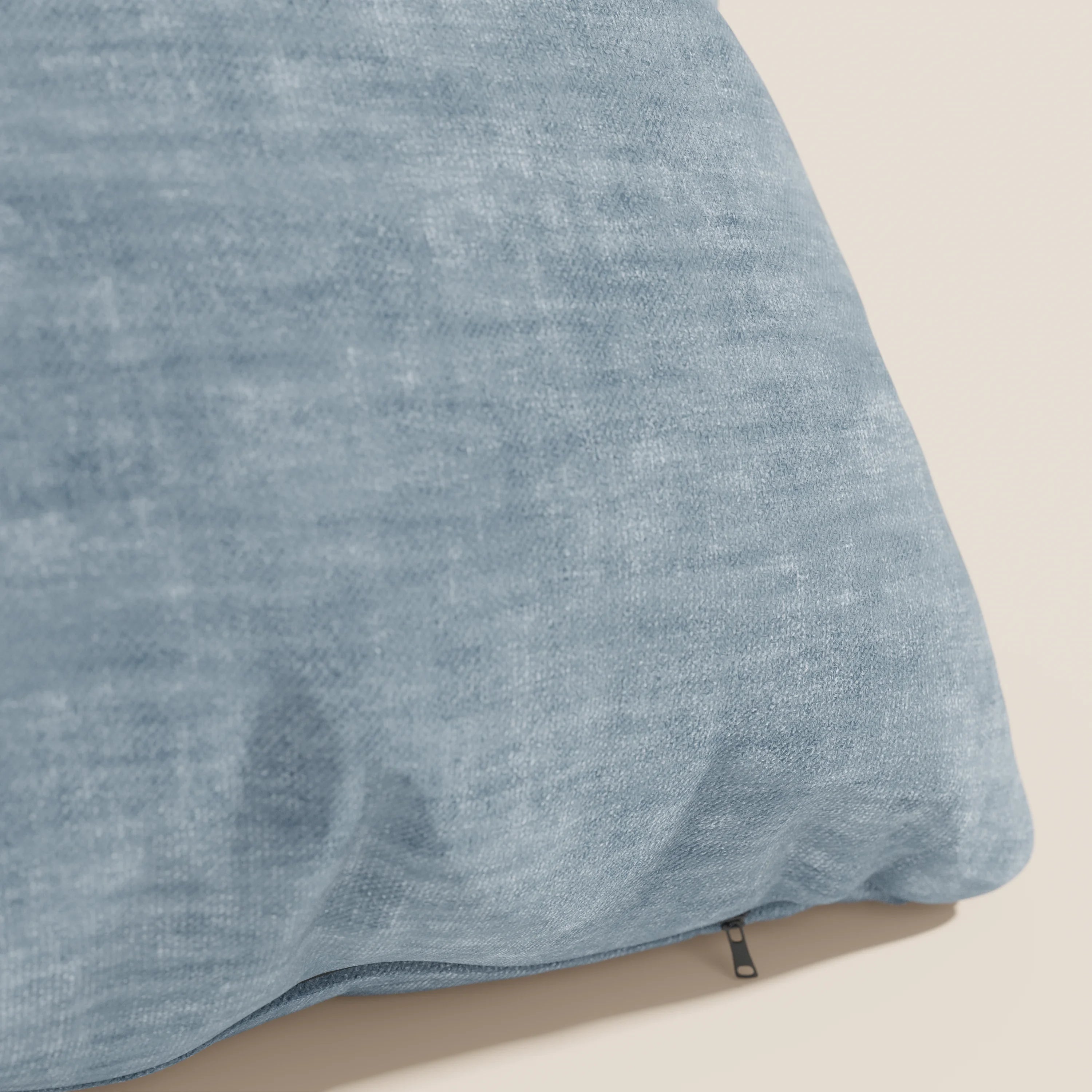 Primo piano del cuscino Sofia di Divani.Store, di colore azzurro, realizzato in morbido velluto Easy Clean T35, antimacchia e impermeabile, con una piccola cerniera nera, su uno sfondo beige chiaro.