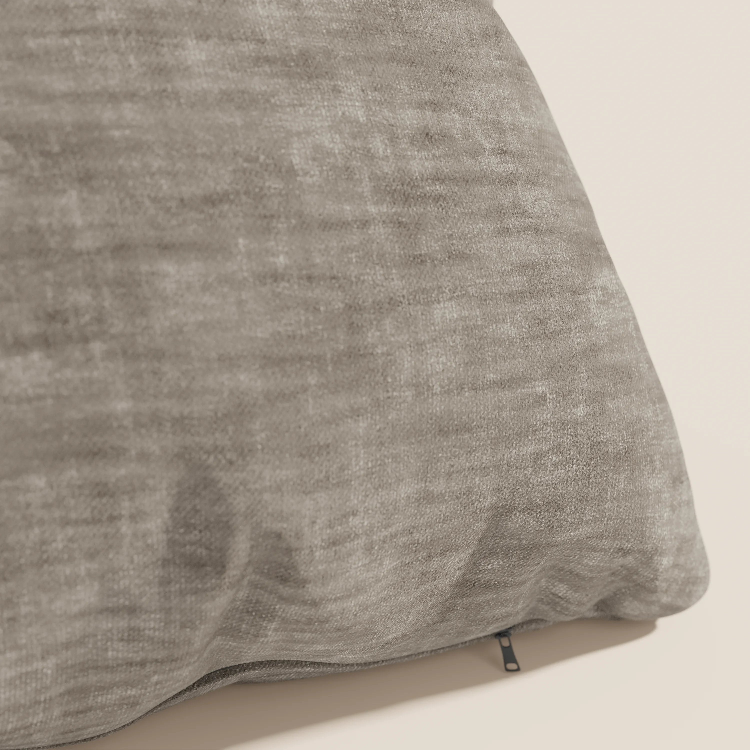 Primo piano del cuscino Sofia di Divani.Store, in grigio. Realizzato in morbido velluto nuvolato (Easy Clean T35), è resistente alle macchie e impermeabile e presenta una cerniera nera. Lo sfondo è semplice e di colore chiaro.