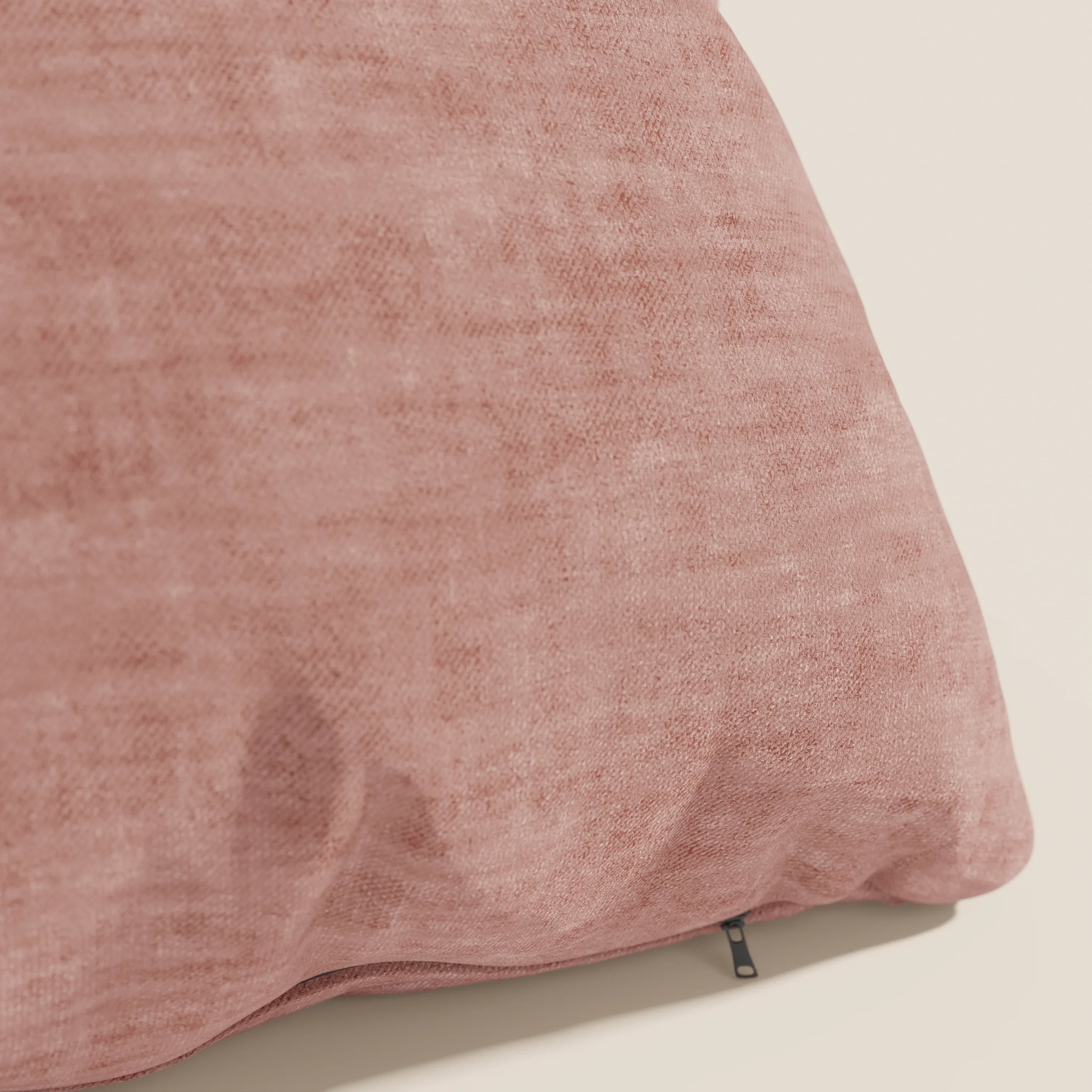 Primo piano di un cuscino Sofia rosa di Divani.Store in morbido velluto nuvolato (Easy Clean T35), resistente alle macchie e all'acqua, con una cerniera a vista. Il cuscino poggia su una superficie chiara su uno sfondo bianco sporco.