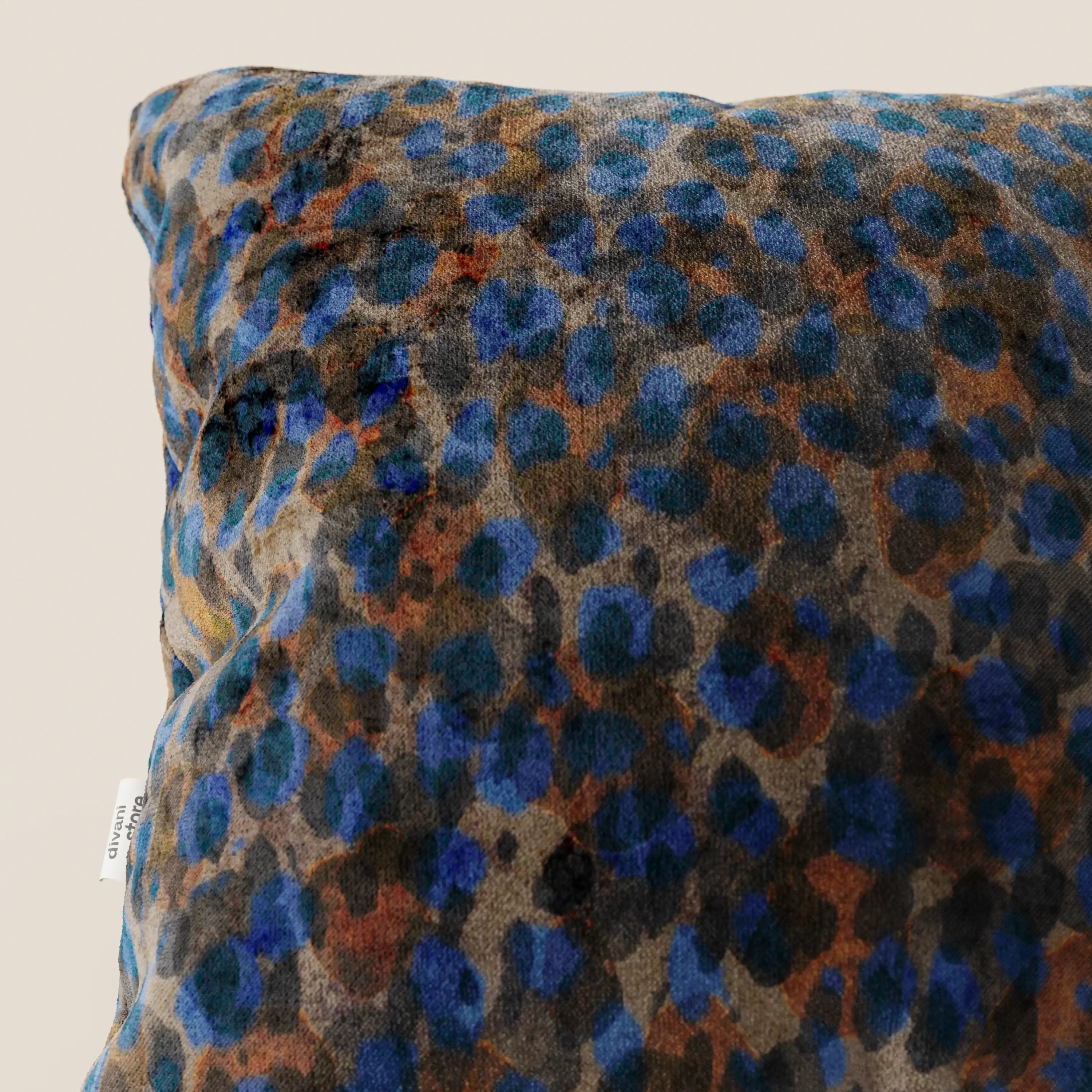Un primo piano dei cuscini decorativi lavabili Divani.Store Brush, realizzati in morbido velluto impermeabile blu, mostra macchie leopardate blu e marroni su sfondo grigio. A sinistra è visibile una piccola etichetta bianca; lo sfondo è beige.