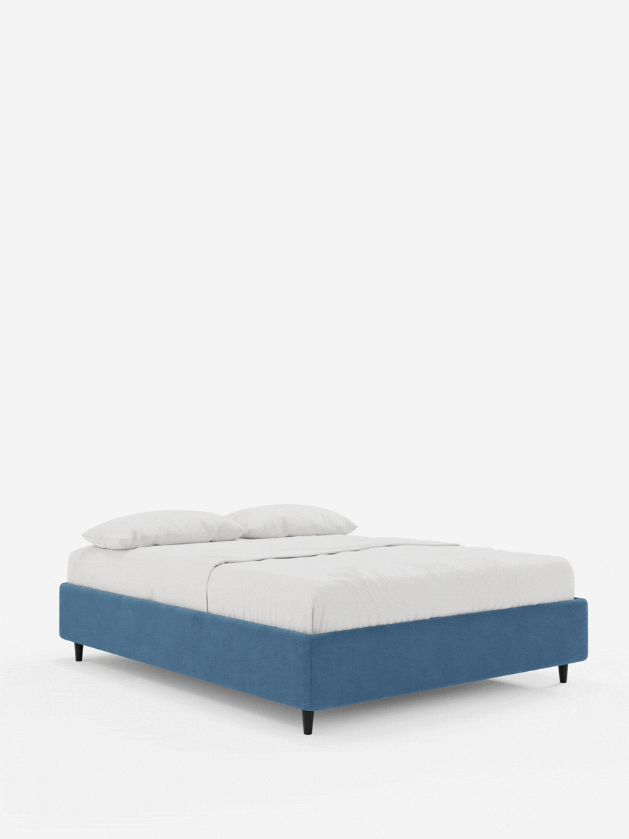 Un letto sommier imbottito ERON con contenitore, rivestito in tessuto impermeabile certificato Oeko-Tex in blu vivace, poggia su quattro piccole gambe affusolate nere; è adornato con lenzuola bianche e due cuscini su uno sfondo chiaro e semplice.
