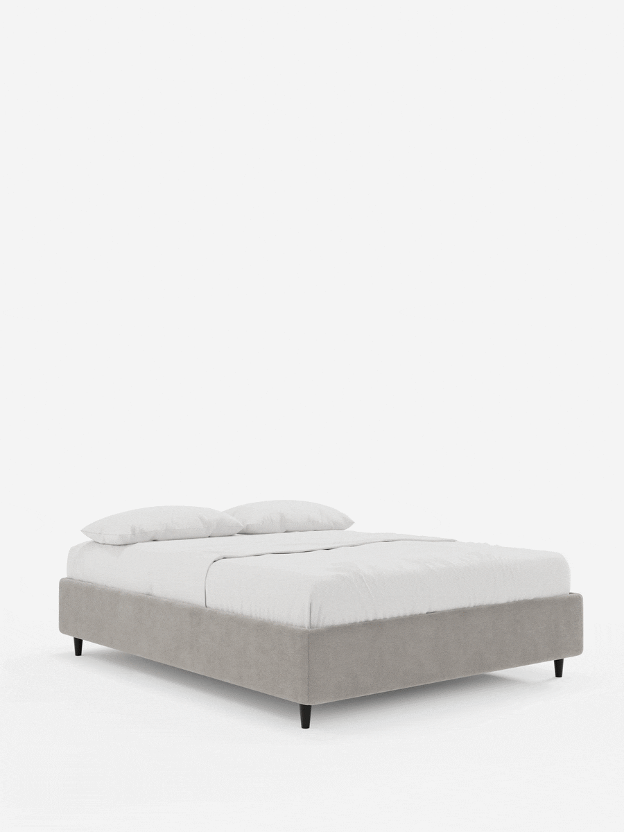 Un letto sommier ERON dal profilo basso, imbottito e dotato di vano contenitore, rivestito in tessuto impermeabile certificato Oeko-Tex color grigio chiaro, con gambe corte e affusolate di colore scuro, rifinito con cura con lenzuola bianche e due cuscini su uno sfondo bianco sporco.