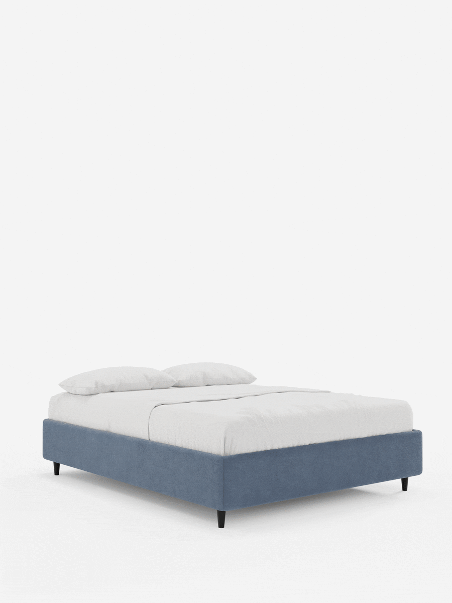 Letto ERON con rete imbottita e contenitore, rivestito in tessuto impermeabile certificato Oeko-Tex di colore blu, con piedini bassi e scuri, rifinito con lenzuola bianche e due cuscini su uno sfondo bianco a tinta unita.