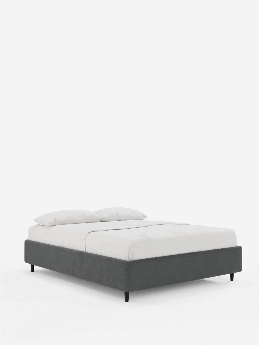 Un letto a piattaforma ERON imbottito con contenitore, realizzato in tessuto impermeabile certificato Oeko-Tex, di colore grigio scuro, poggia su corte gambe nere su uno sfondo bianco, rivestito con lenzuola bianche immacolate e due cuscini coordinati.