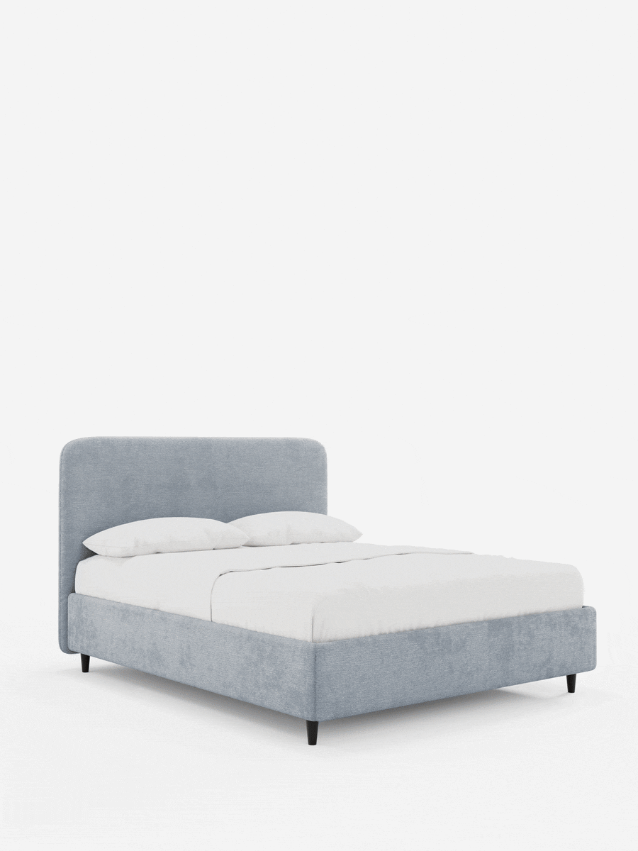 Un letto KAELOS minimal imbottito – con contenitore, gambe alte e testiera arrotondata in tessuto ciniglia azzurro – spicca su uno sfondo bianco, caratterizzato da una testiera arrotondata, gambe sottili e affusolate e una raffinata biancheria da letto bianca.