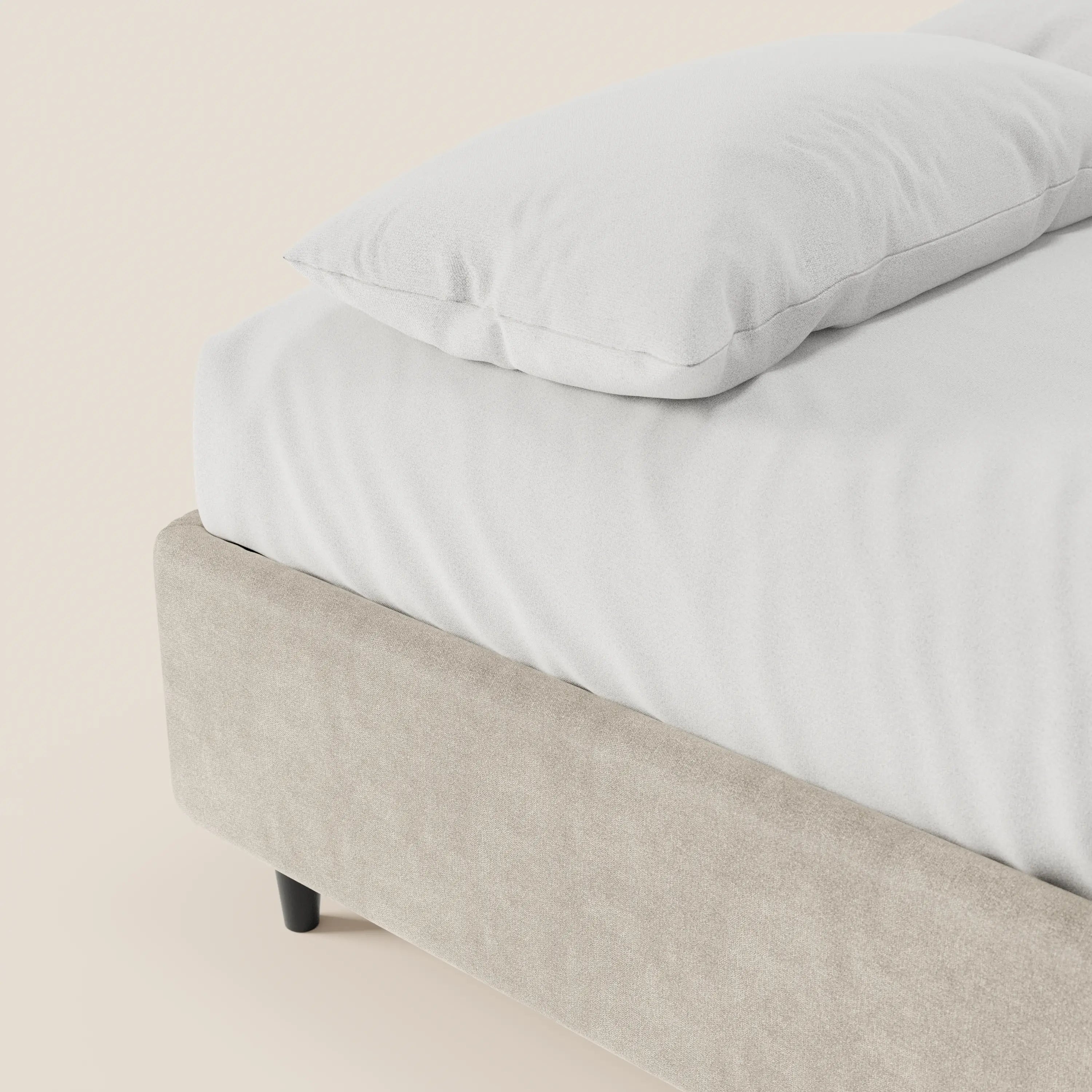 Un primo piano del letto sommier imbottito ERON di Letti.store mette in risalto il suo tessuto beige chiaro impermeabile e certificato Oeko-Tex, un morbido cuscino bianco, un lenzuolo con angoli liscio e le gambe corte nere, il tutto su uno sfondo color crema tenue.