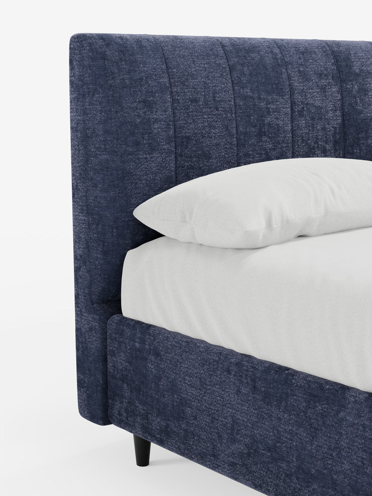 Un primo piano del letto imbottito ONYX con contenitore e testiera verticale in tessuto certificato Oeko-Tex® di Letti.store ne mette in risalto il tessuto da rivestimento blu scuro. Il letto presenta una testiera imbottita a pannelli verticali, un cuscino bianco spesso e biancheria da letto bianca abbinata. Di questo letto imbottito contenitore sono visibili solo un angolo e un lato, con le corte gambe nere. Lo sfondo è bianco a tinta unita.