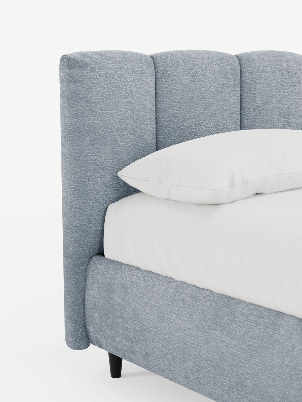 Un primo piano del letto imbottito ARKAM con contenitore e testiera a lavorazione verticale, caratterizzato da una testiera imbottita blu chiaro con texture e cuciture verticali, un cuscino bianco e gambe cilindriche scure su sfondo bianco.