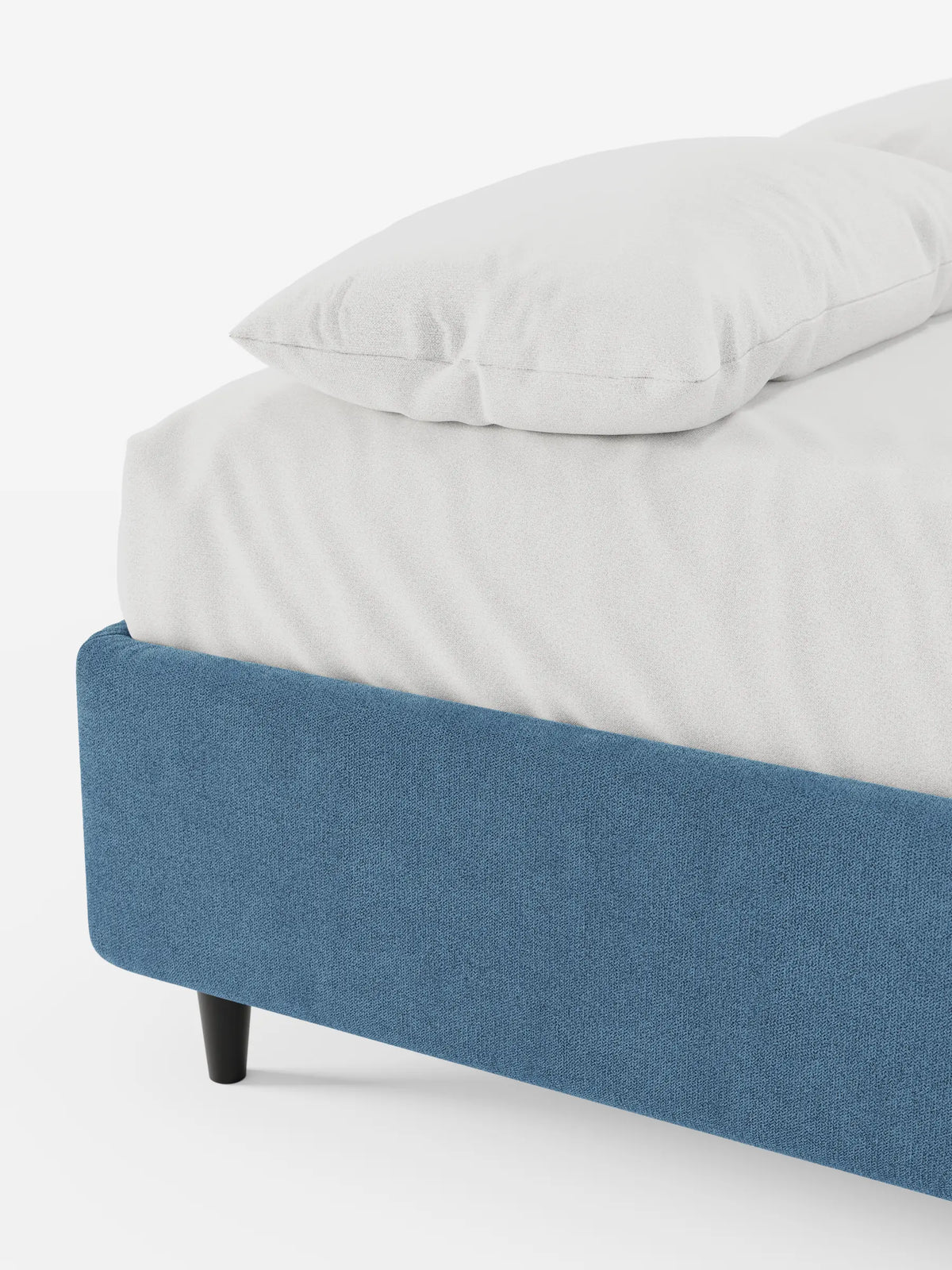 Un primo piano dell'angolo di un letto sommier imbottito ERON con vano contenitore in tessuto impermeabile certificato Oeko-Tex, in tessuto blu testurizzato, con un lenzuolo bianco e un cuscino appoggiati su una gamba corta e scura, su uno sfondo bianco.