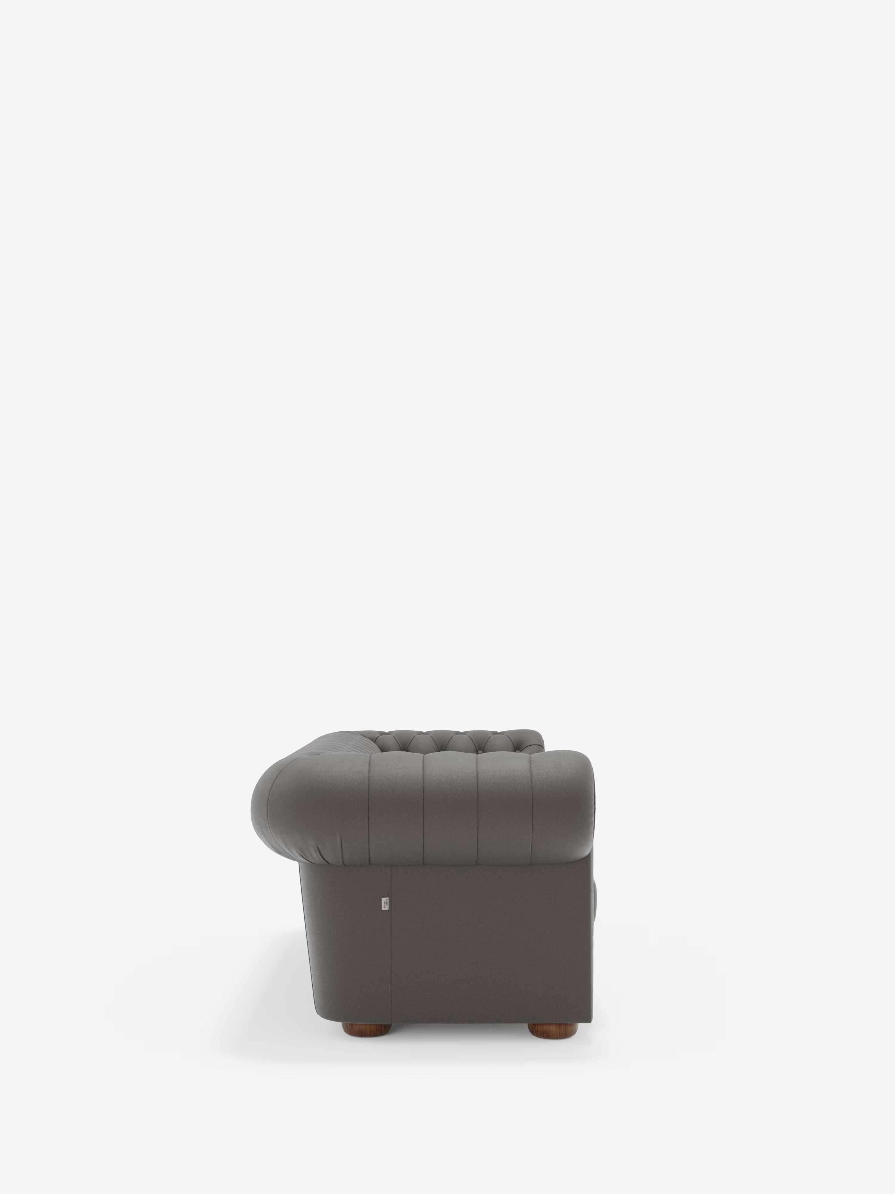 Una vista laterale della “Chester – Divano Chesterfield in vera pelle spessorata a 2 e 3 posti” di Divani.Store, una poltrona imbottita di colore grigio scuro, con rivestimento trapuntato, braccioli ricurvi e piedini in legno, che richiama il classico stile Chesterfield e si stacca su uno sfondo bianco a tinta unita.