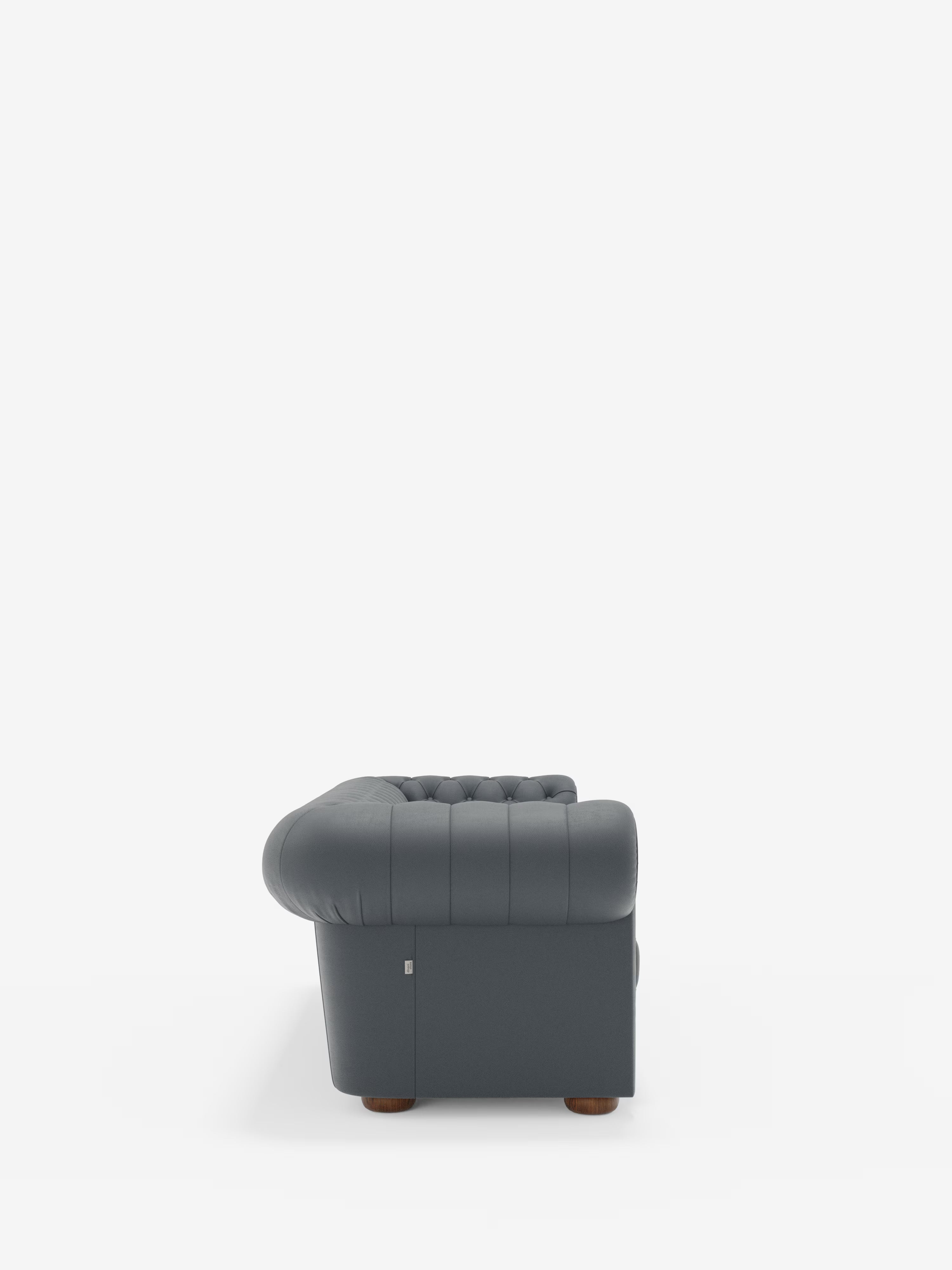 Vista laterale del Chester 4 Posti 3 sedute in Vera Pelle – Chesterfield Capitonné Artigianale di Divani.Store, una poltrona imbottita in vera pelle di colore grigio scuro con rivestimento capitonné, braccioli e schienale arrotondati, gambe in legno e una piccola etichetta metallica nella parte inferiore sinistra — che richiama il classico design Chesterfield — su uno sfondo bianco a tinta unita.