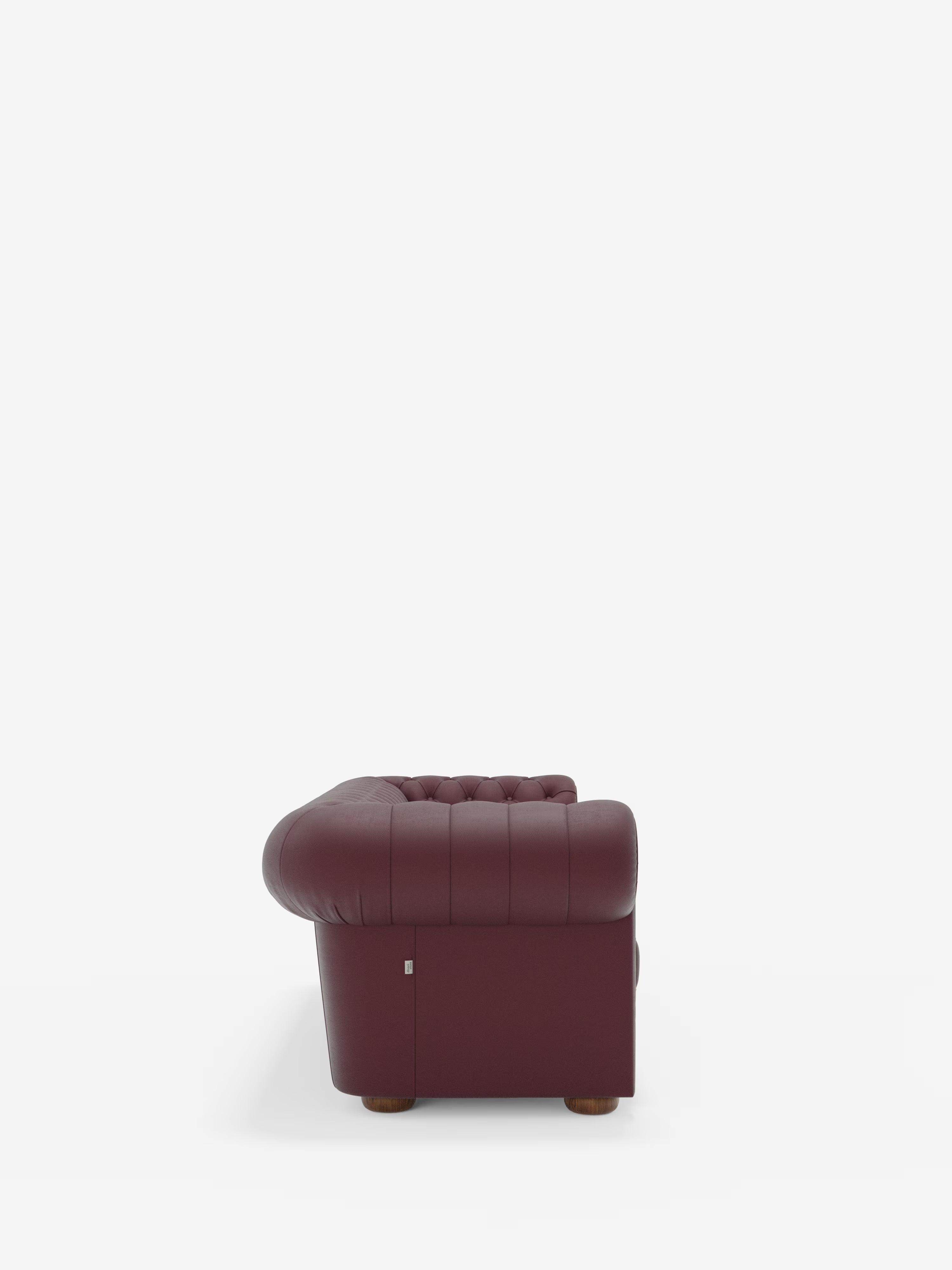 Vista laterale del divano Chester 4 Posti 3 sedute in Vera Pelle – Chesterfield Capitonné Artigianale di Divani.Store, caratterizzato da rivestimento in vera pelle capitonné color bordeaux, braccioli e schienale leggermente arrotondati, piedini in legno e una discreta etichetta metallica vicino alla base. Presentato su uno sfondo bianco a tinta unita per mettere in risalto la silhouette e la trama artigianale del rivestimento.