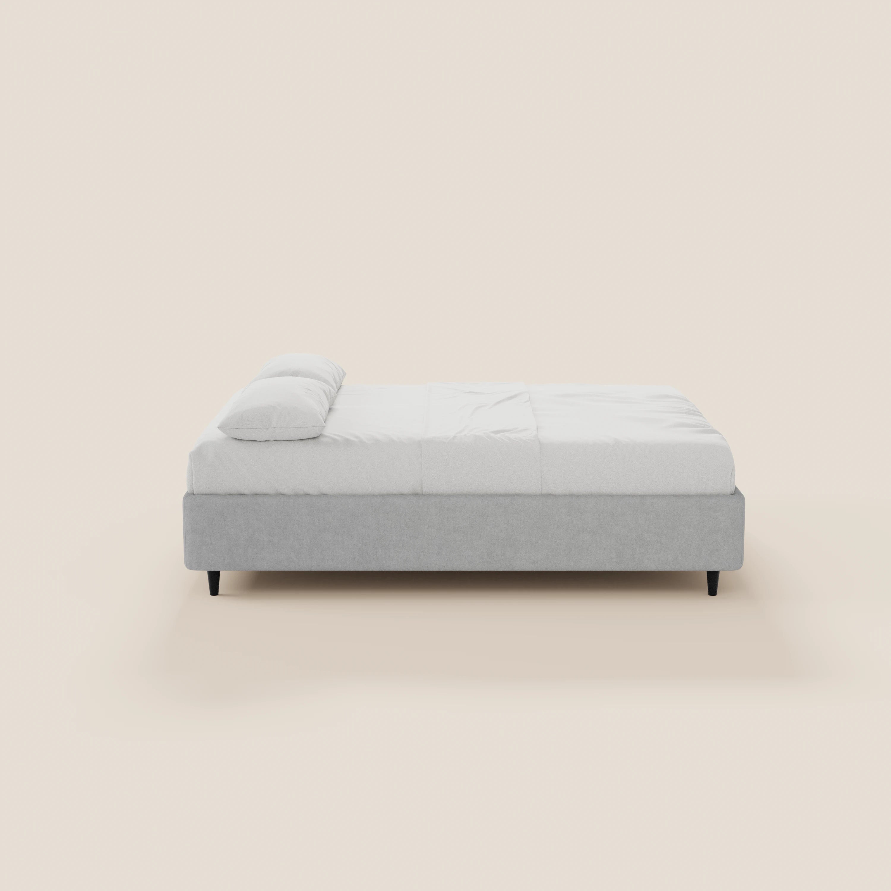 Il letto sommier ERON di Letti.store presenta un design moderno e minimalista, con una struttura imbottita impermeabile Oeko-Tex di colore grigio chiaro, gambe nere, materasso e biancheria da letto bianchi, ed è raffigurato di profilo su uno sfondo bianco sporco.