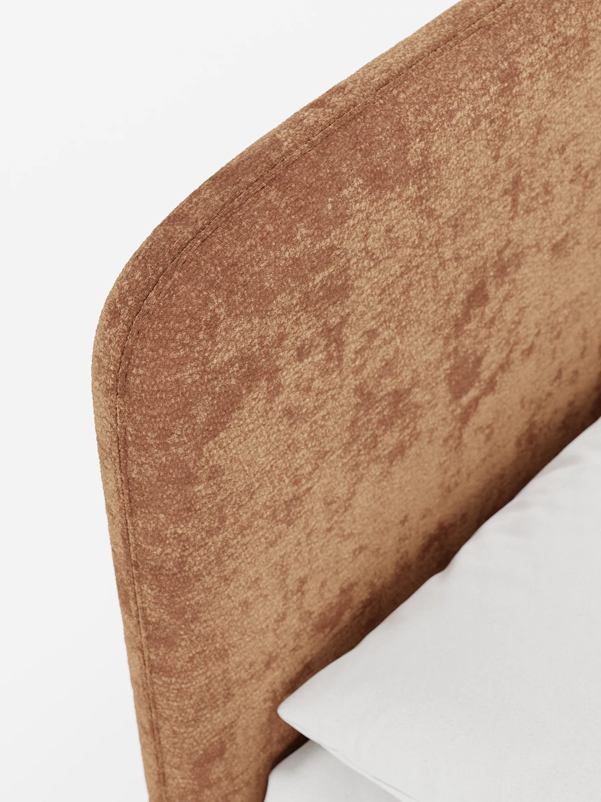 Un primo piano della testiera imbottita KAELOS “letto minimal”, dalla forma arrotondata e rivestita in tessuto color terracotta con effetto screziato e texture, con un cuscino bianco a tinta unita appoggiato contro di essa su uno sfondo chiaro.