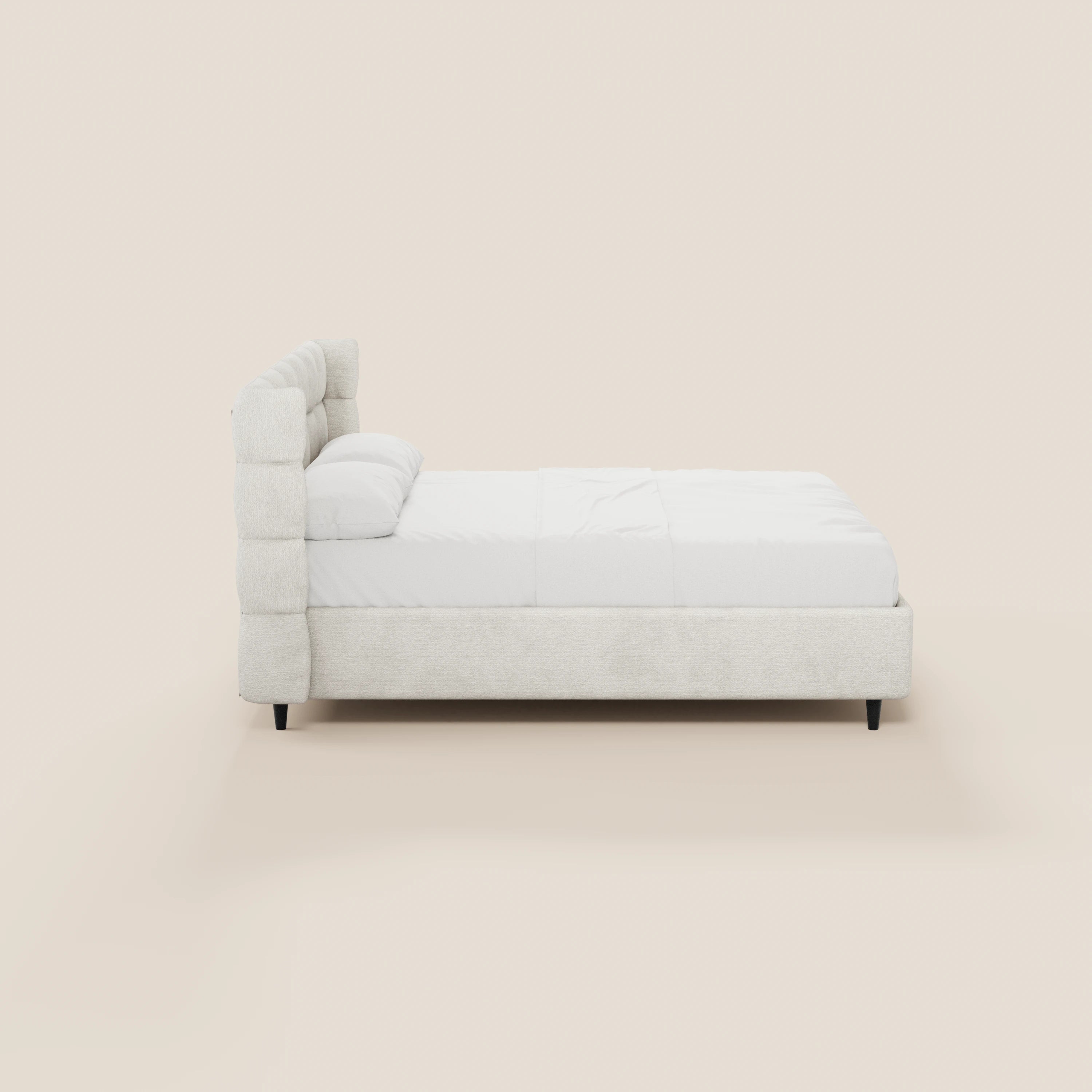 Vista laterale del letto imbottito RAGNAR con contenitore e testiera capitonné avvolgente di Letti.store, in tessuto bianco sporco certificato Oeko-Tex®, caratterizzato da una struttura a profilo ribassato, due cuscini, un lenzuolo bianco e gambe corte scure su uno sfondo beige.