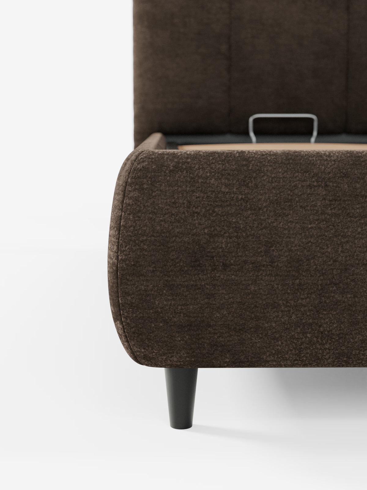 Un primo piano dell'angolo del letto imbottito ARKAM con contenitore e testiera a lavorazione verticale di Letti.store, caratterizzato da un rivestimento in tessuto marrone scuro certificato Oeko-Tex®, bordi arrotondati, una gamba affusolata nera e una maniglia metallica a vista sulla base del letto. Lo sfondo è bianco liscio.
