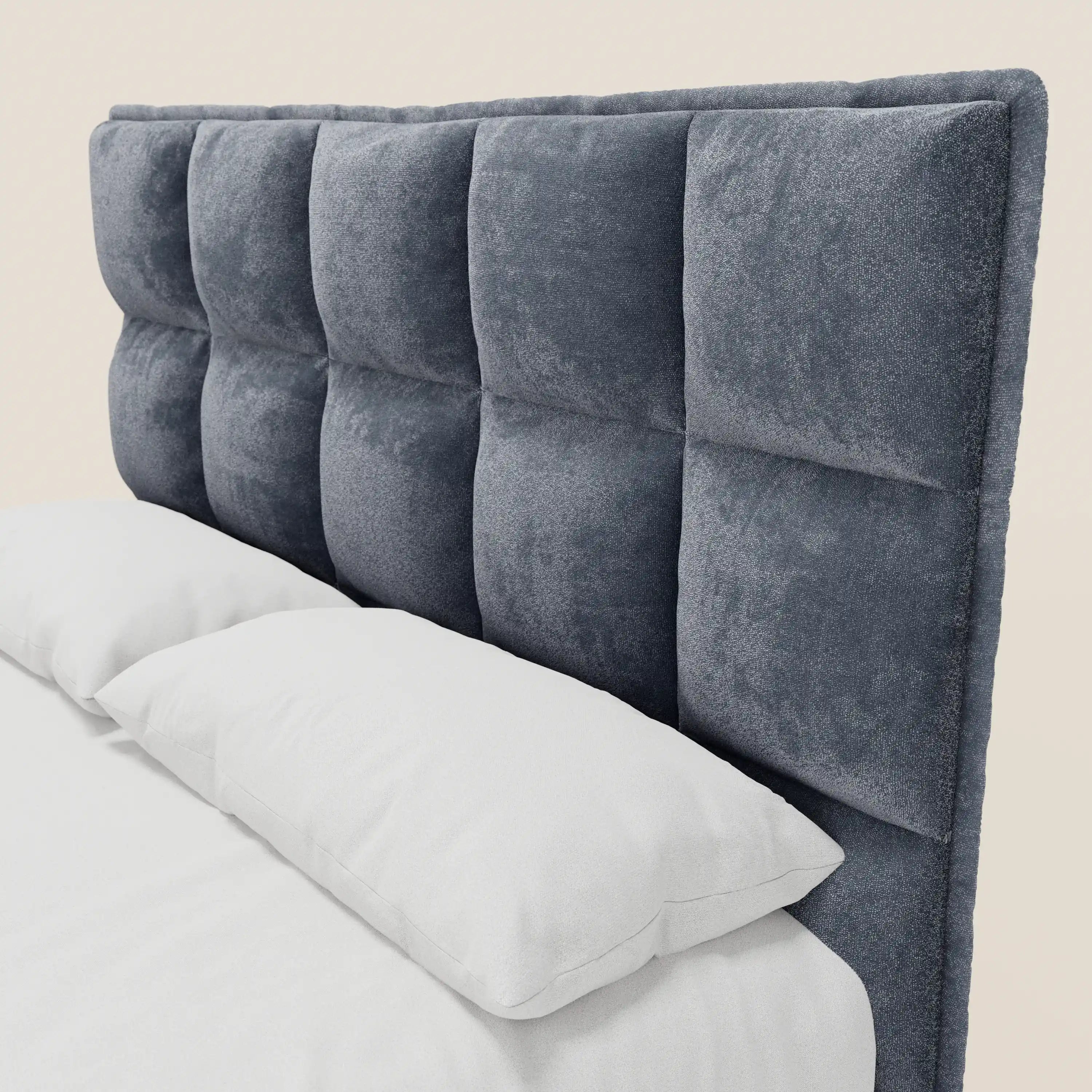 Primo piano del letto imbottito AURO di Letti.store, caratterizzato da una testiera alta e capitonné con motivo a quadrati, realizzata in ciniglia grigio scuro certificata Oeko-Tex®. Due cuscini bianchi poggiano contro la testiera su un materasso bianco.