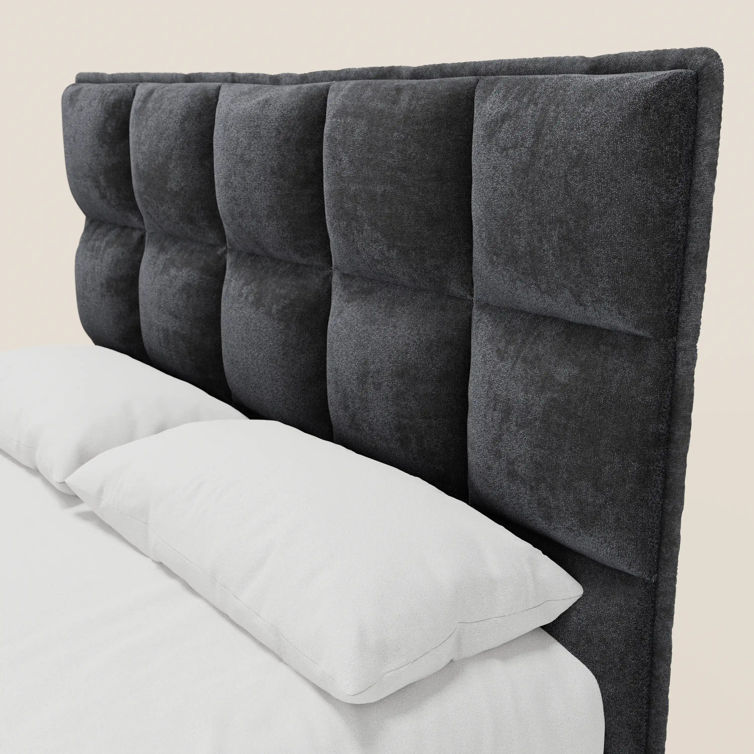 Un primo piano del letto AURO di Letti.store mette in risalto la sua testiera alta e capitonné, realizzata in ciniglia grigio scuro certificata Oeko-Tex®. La biancheria da letto di un bianco brillante crea un contrasto con la testiera dalla trama rigata, che si staglia su uno sfondo beige chiaro.