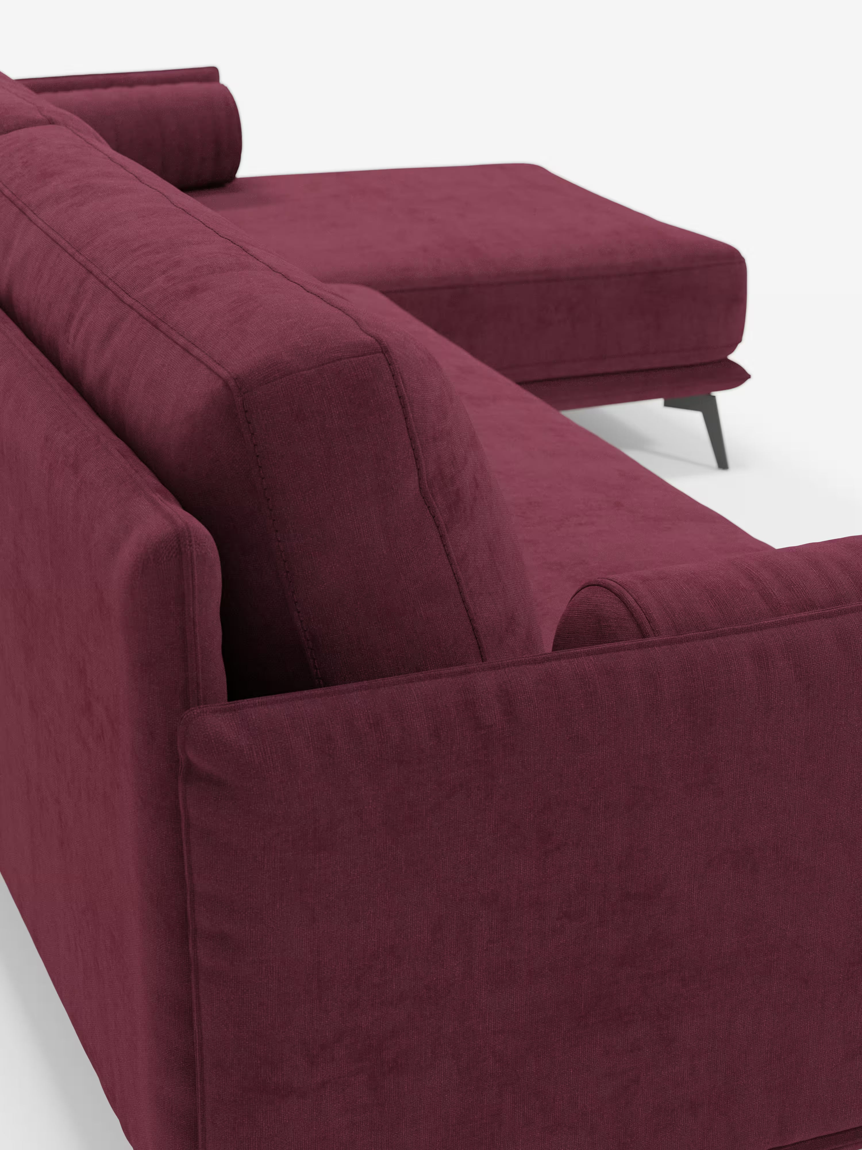 Il divano moderno angolare con penisola Basalto di Divani.Store è presentato in una vista posteriore angolata, caratterizzato da un tessuto T31 color bordeaux intenso e resistente alle macchie. Questo divano componibile contemporaneo si distingue per le linee pulite, i cuscini spessi imbottiti di piume, la sezione chaise longue, i cuscini cilindrici e le slanciate gambe in metallo nero, il tutto su uno sfondo bianco a tinta unita.