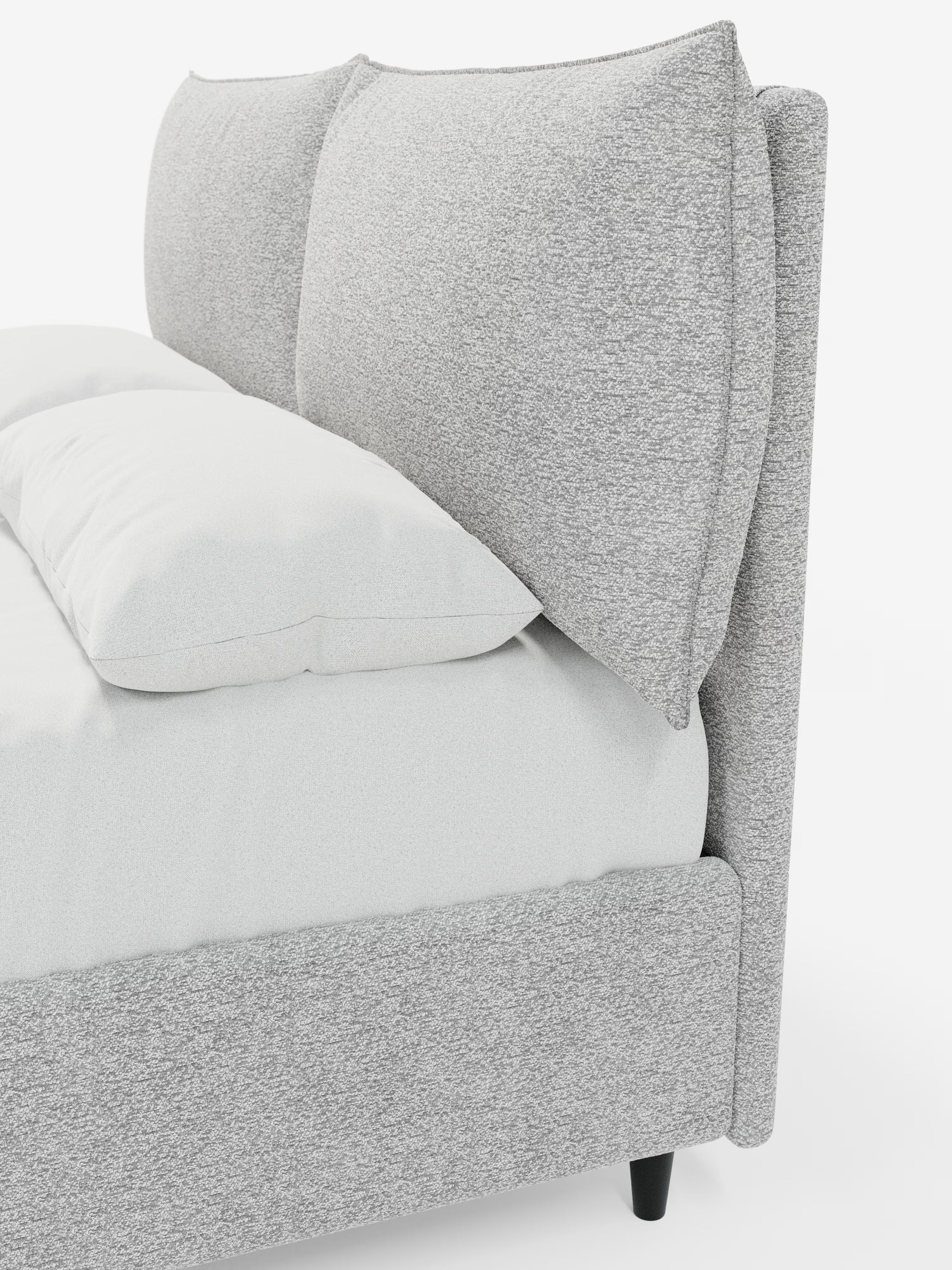 Un primo piano del letto imbottito ZAYRON di Letti.store mette in risalto la struttura rivestita in tessuto grigio chiaro certificato Oeko-Tex® e i cuscini della testiera oversize e regolabili, simili a cuscini da letto. Il letto è rivestito con biancheria da letto di un bianco brillante e poggia su gambe corte nere, creando un look minimalista e contemporaneo.