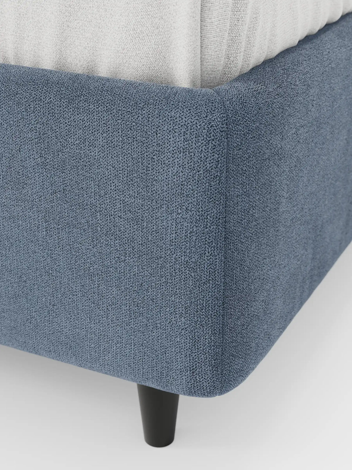 Primo piano di un letto sommier imbottito ERON color blu polveroso, dotato di vano contenitore in tessuto impermeabile certificato Oeko-Tex con angoli arrotondati, poggiante su una gamba cilindrica scura e sormontato da un materasso neutro di colore chiaro.
