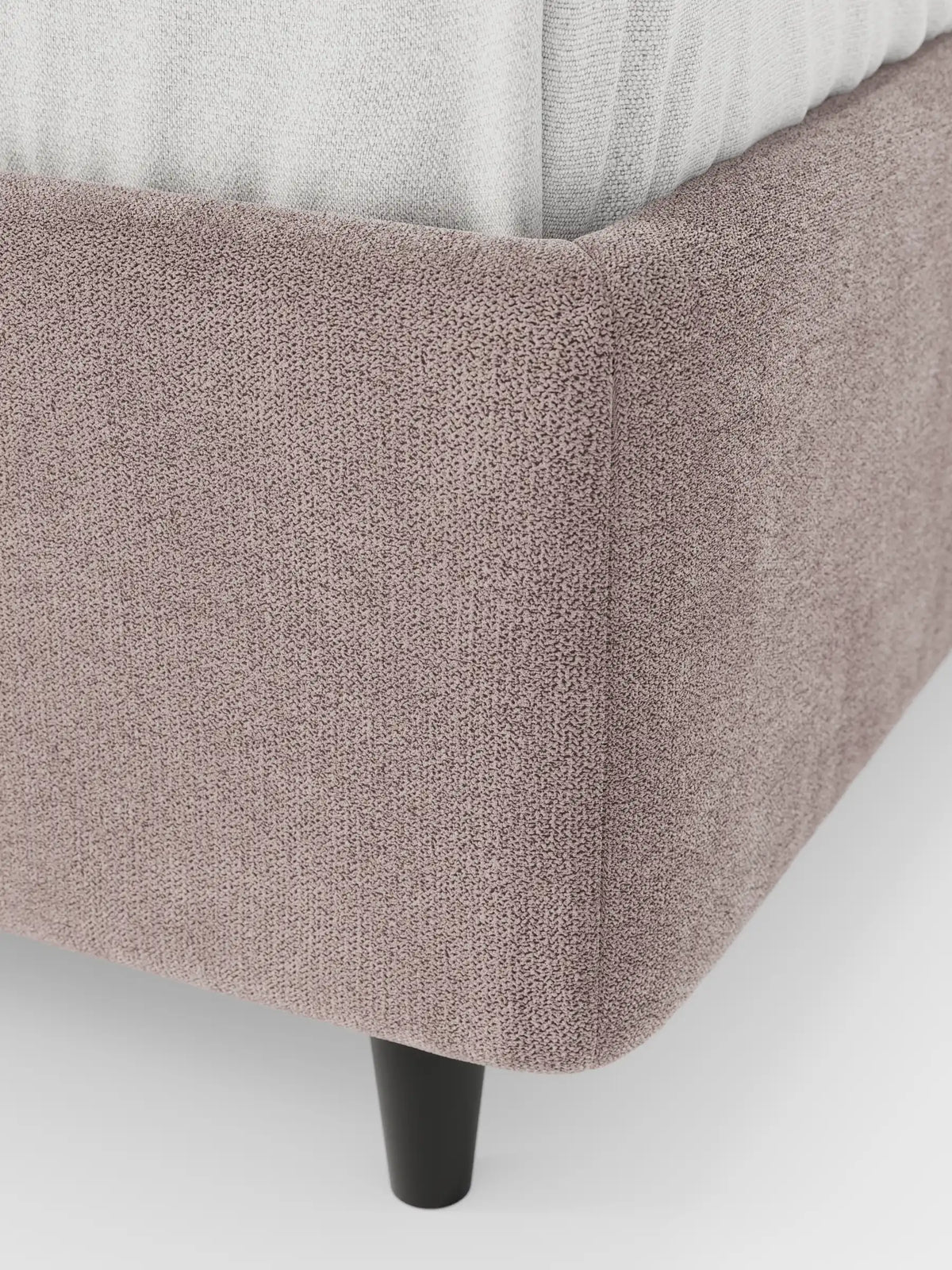 Un primo piano del letto sommier imbottito ERON con contenitore in tessuto impermeabile certificato Oeko-Tex, caratterizzato da un angolo imbottito color malva con bordo arrotondato, una gamba cilindrica nera e biancheria da letto chiara sopra.