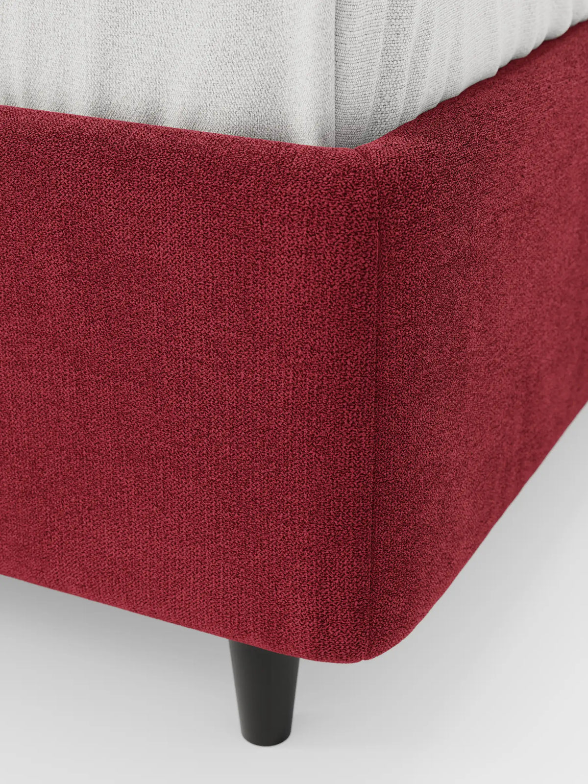 Un primo piano di un letto sommier ERON imbottito, di colore rosso intenso e dalla superficie ruvida, dotato di vano contenitore in tessuto impermeabile certificato Oeko-Tex; si notano una semplice gamba nera e il bordo di un materasso di colore chiaro su uno sfondo bianco a tinta unita.