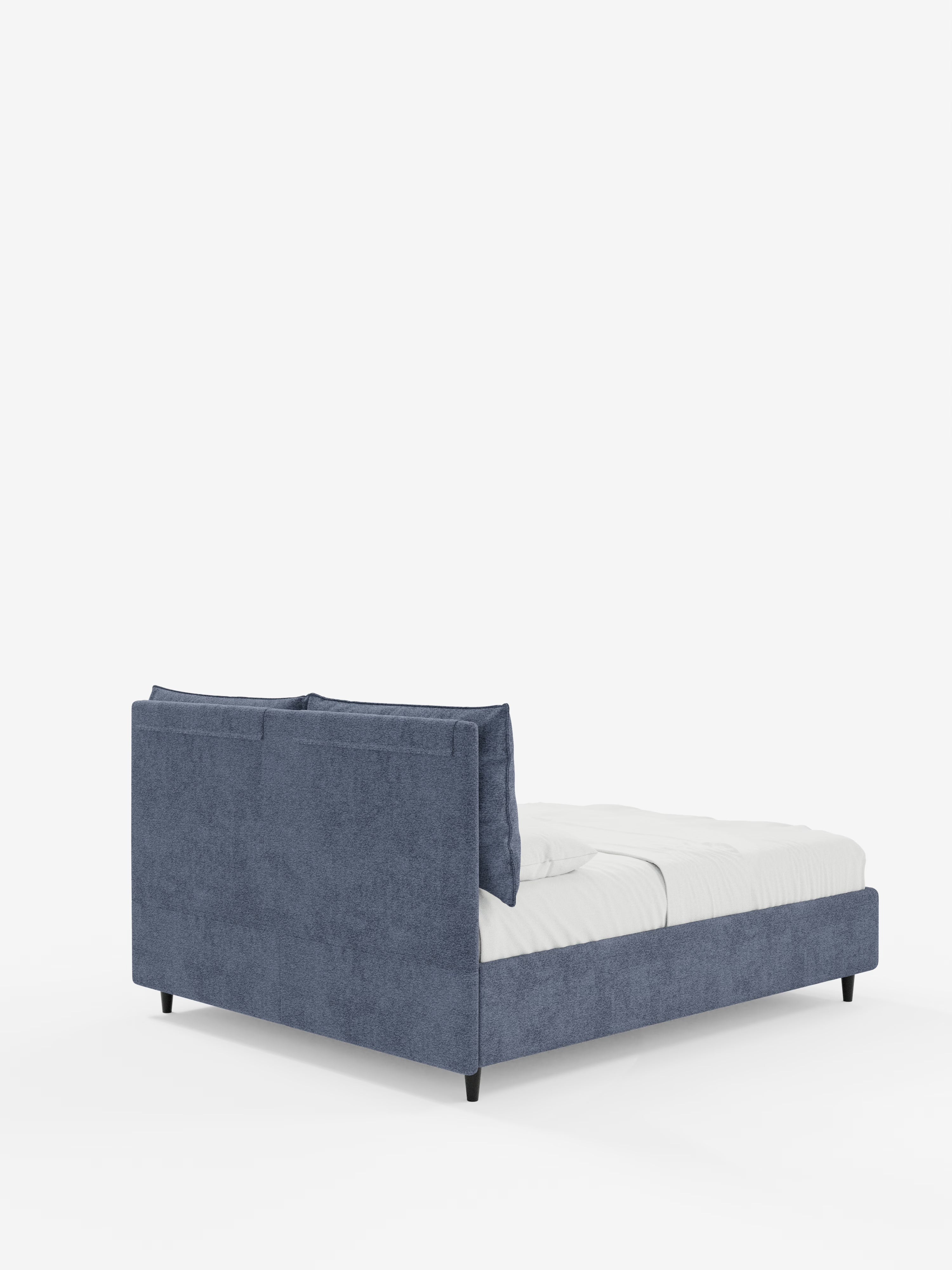 Il moderno letto imbottito ZAYRON con contenitore e morbidi cuscini regolabili di Letti.store è presentato con struttura e testiera imbottite di colore blu, certificate Oeko-Tex®, materasso bianco e biancheria da letto. Il letto poggia su corte gambe nere su uno sfondo bianco a tinta unita; l’angolazione della ripresa mette in risalto la parte posteriore e una porzione laterale del mobile.