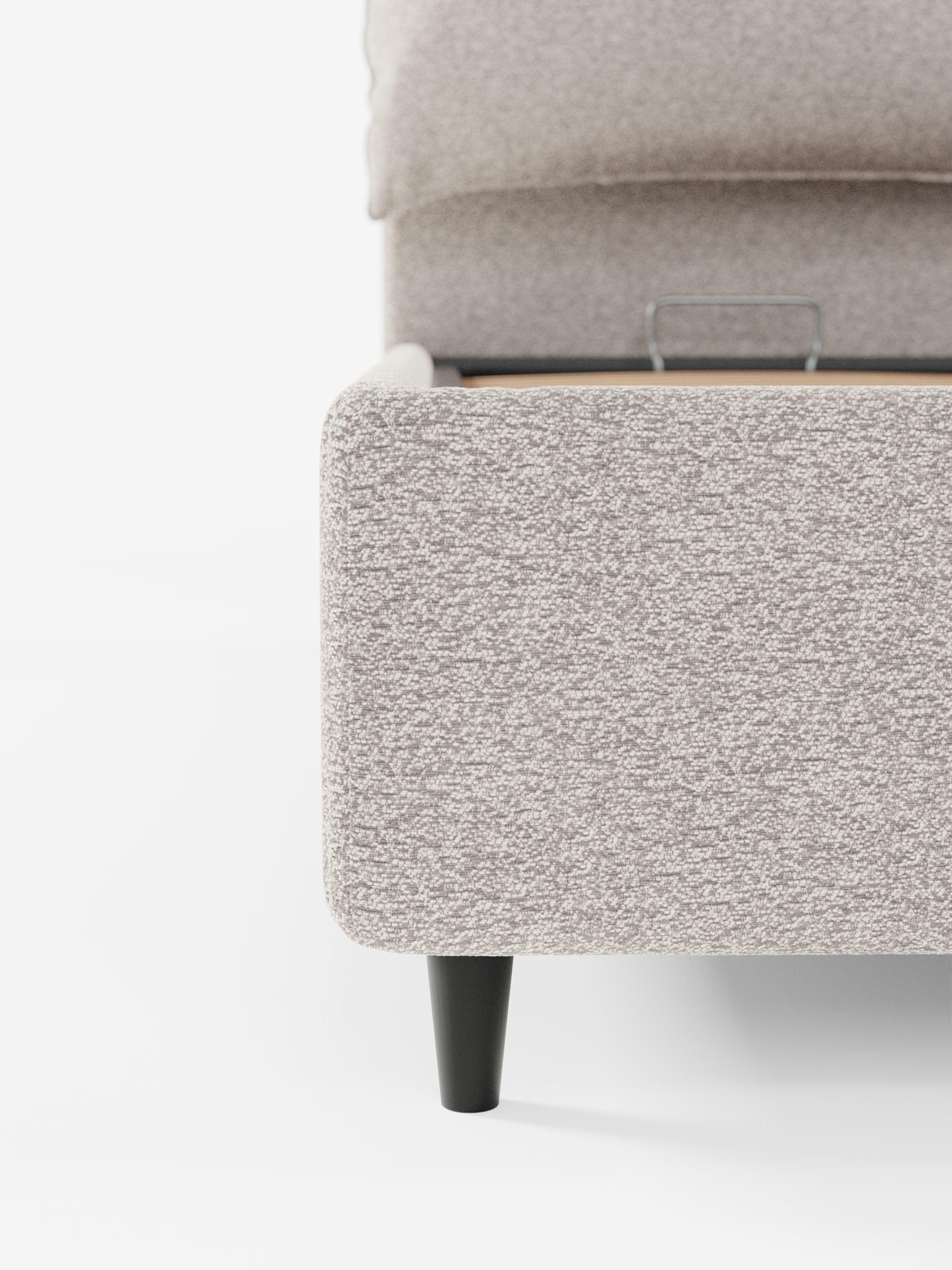 Primo piano della struttura del letto imbottito ZAYRON di Letti.store, realizzata in tessuto beige testurizzato certificato Oeko-Tex® dal design moderno. L’immagine mette in risalto l’angolo inferiore e una gamba affusolata nera, su sfondo bianco. Una maniglia in metallo all’interno della struttura indica la presenza di un vano contenitore sotto il letto.