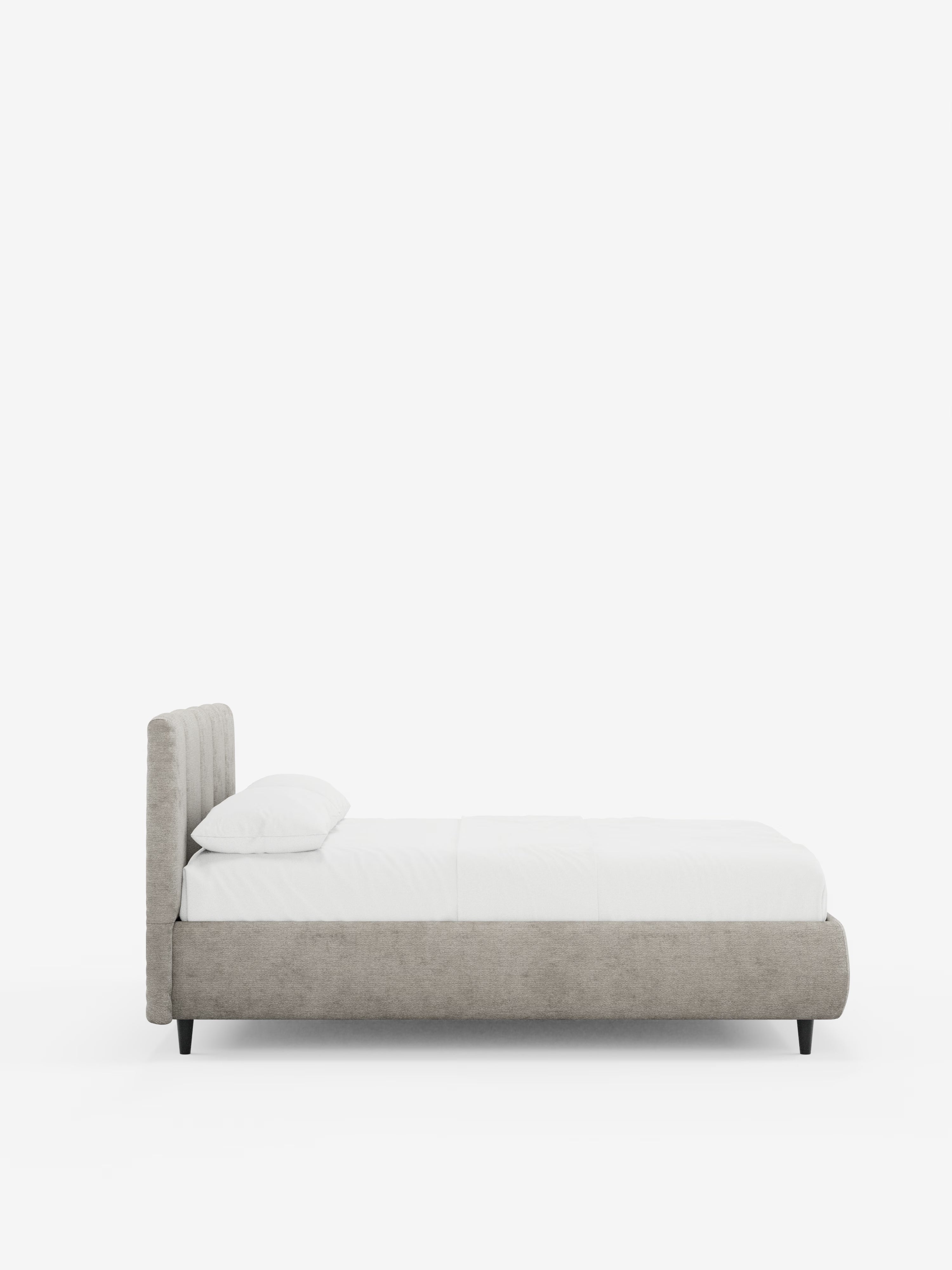 Vista laterale del letto imbottito ARKAM con contenitore e testiera a lavorazione verticale di Letti.store, caratterizzato da una testiera imbottita grigio chiaro certificata Oeko-Tex® e da una struttura in stile shabby chic. Il letto è adornato da un cuscino bianco e da biancheria da letto bianca, presenta gambe corte nere ed è posizionato su uno sfondo bianco a tinta unita.