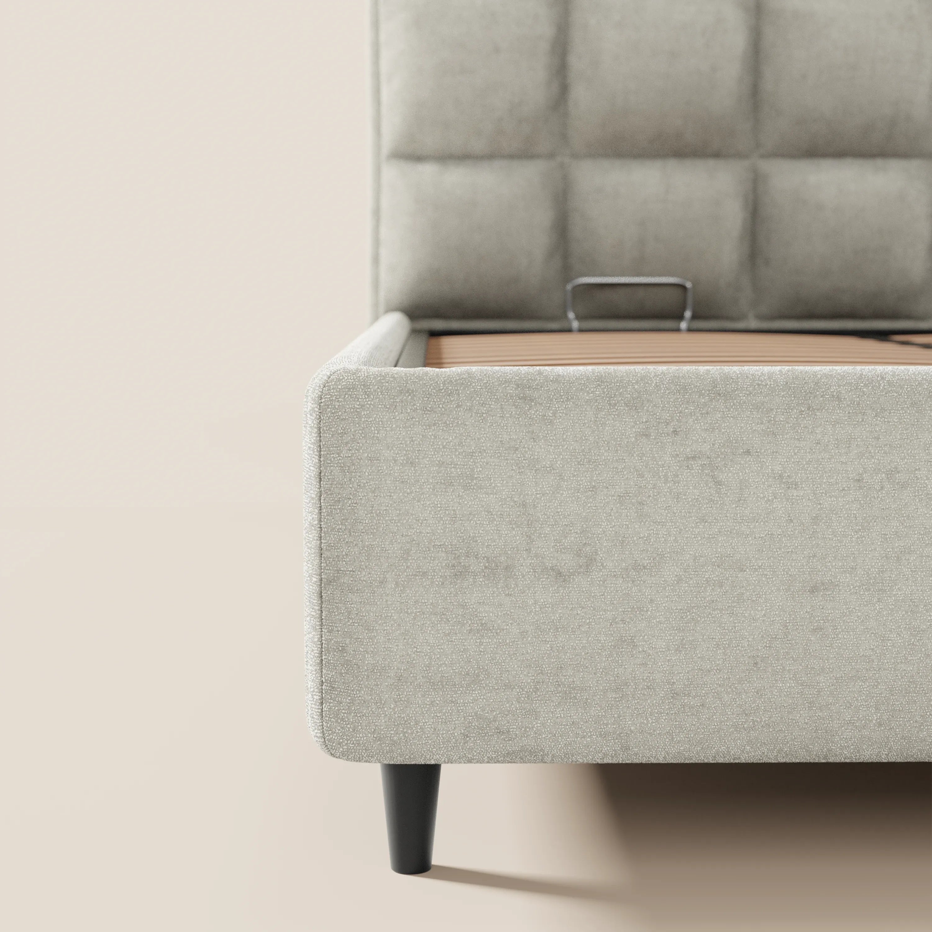 Un primo piano del letto imbottito AURO di Letti.store, realizzato in morbida ciniglia beige certificata Oeko-Tex®, che mostra l'angolo della pediera, le gambe alte in legno scuro, le doghe di sostegno a vista e parte della sua alta testiera capitonné, su uno sfondo di parete beige chiaro. Materasso non incluso.