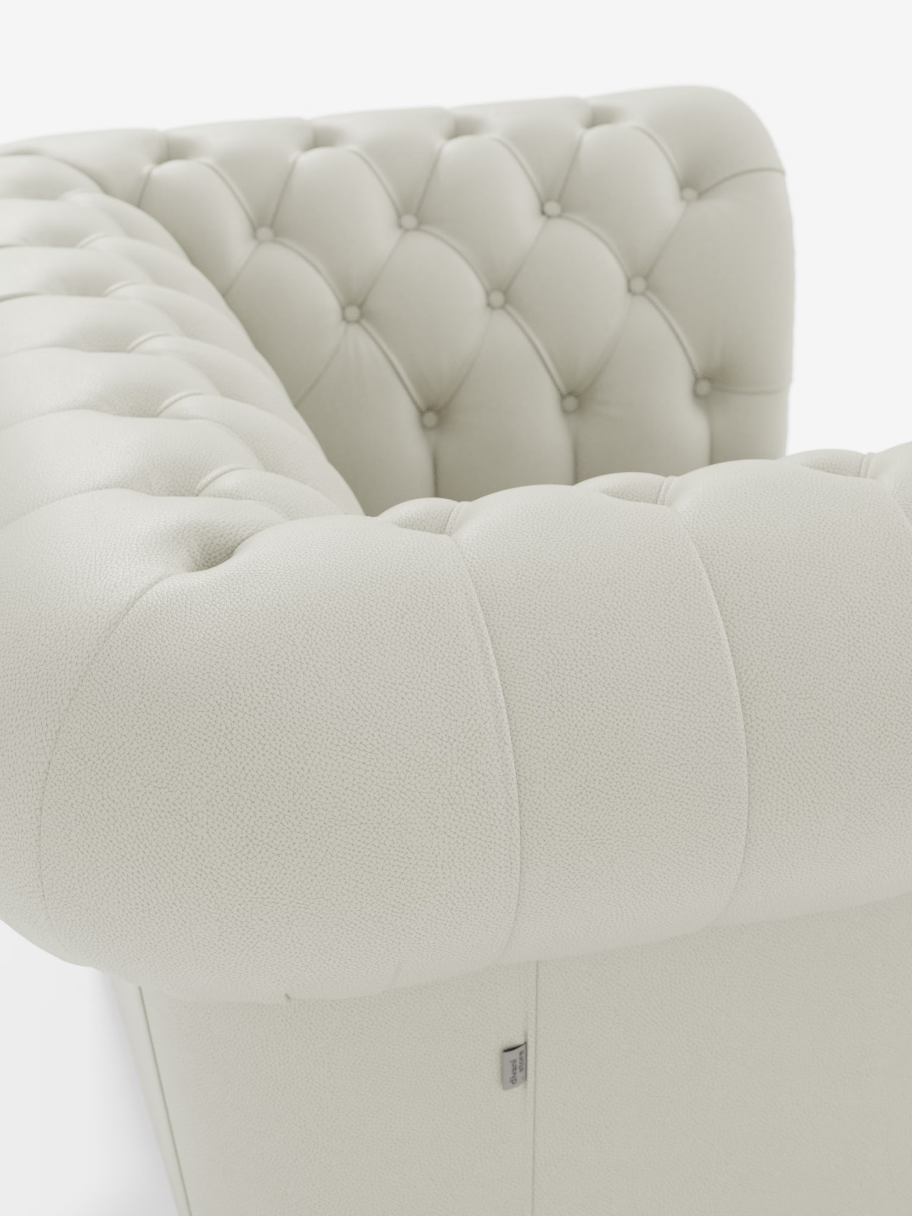 Primo piano della poltrona Chester in vera pelle spessorata con capitonné artigianale di Divani.Store, caratterizzata da un rivestimento in vera pelle color crema con capitonné, dettagli artigianali a bottoni e braccioli arrotondati. La superficie ruvida e una piccola etichetta metallica sul lato risaltano sullo sfondo bianco a tinta unita.