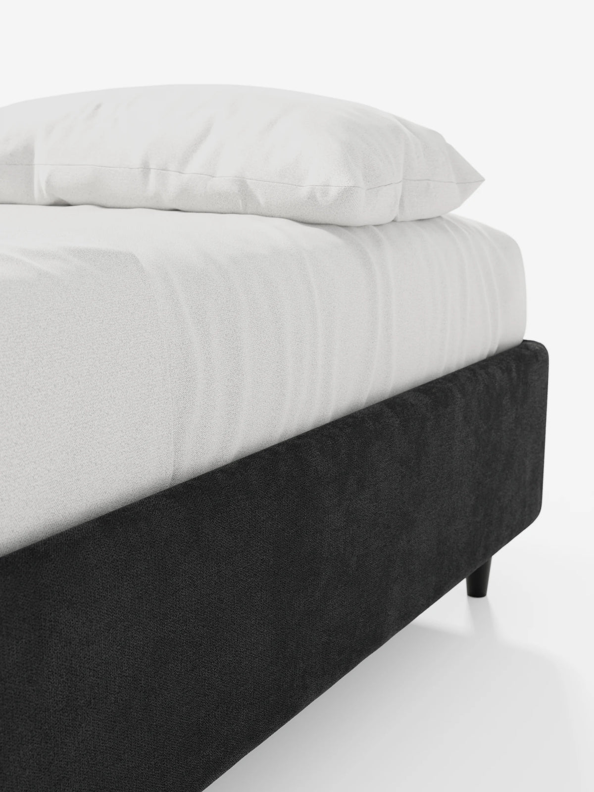 Primo piano di un letto sommier imbottito ERON con contenitore, rivestito in tessuto impermeabile certificato Oeko-Tex di colore grigio scuro, con gambe affusolate nere, un morbido materasso bianco e un cuscino bianco su uno sfondo chiaro e uniforme.