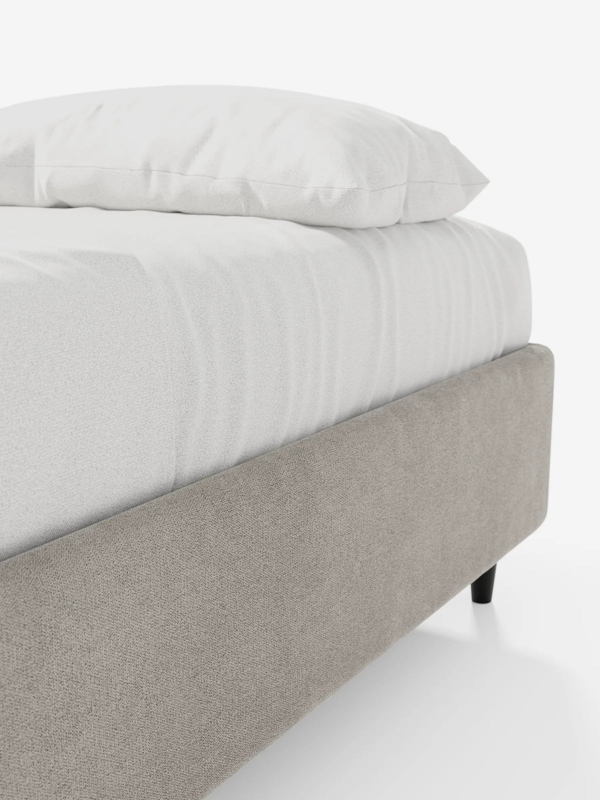 Una vista ravvicinata da un angolo di un letto sommier imbottito ERON con vano contenitore in tessuto impermeabile certificato Oeko-Tex color taupe, con gambe affusolate scure, su cui poggiano un materasso bianco candido e un cuscino singolo, su uno sfondo chiaro e semplice.