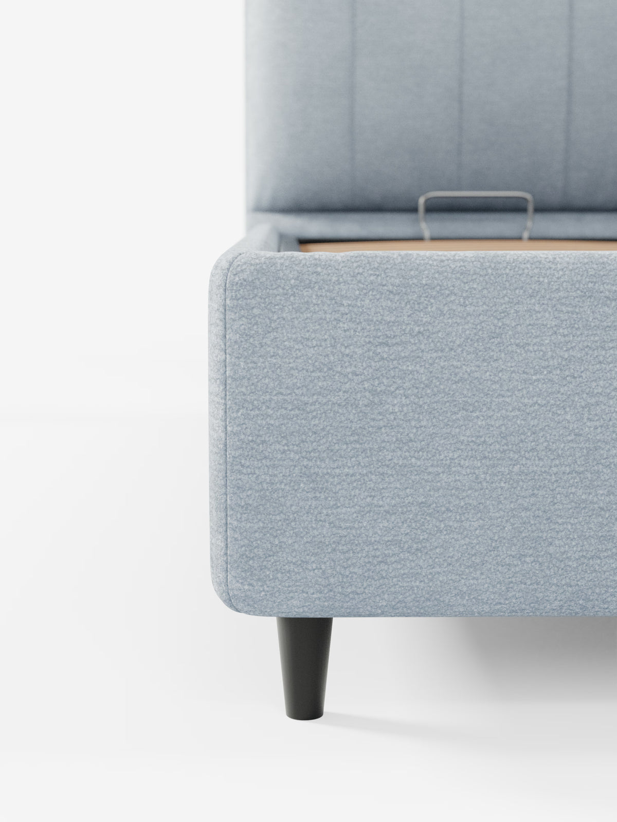 Primo piano dell'angolo del letto imbottito ONYX con vano contenitore e testiera verticale in tessuto certificato Oeko-Tex® di Letti.store, caratterizzato da una struttura imbottita azzurra con gambe affusolate nere. La testiera imbottita e la pediera sono visibili su uno sfondo bianco a tinta unita, mentre all’interno del vano contenitore del letto si intravede parzialmente una maniglia in metallo.