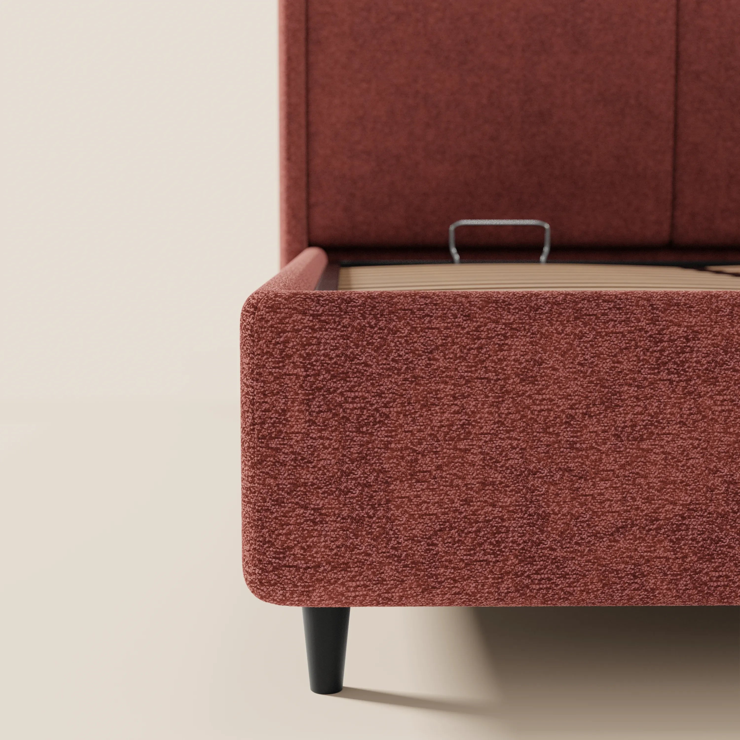 Primo piano del letto di design VALKIRIA di Letti.store, con rivestimento in tessuto color ruggine, gambe affusolate nere e maniglia in metallo a vista sulle doghe in legno. Dotato di vano contenitore e testiera avvolgente con 2 cuscini imbottiti. Sfondo neutro a tinta unita.
