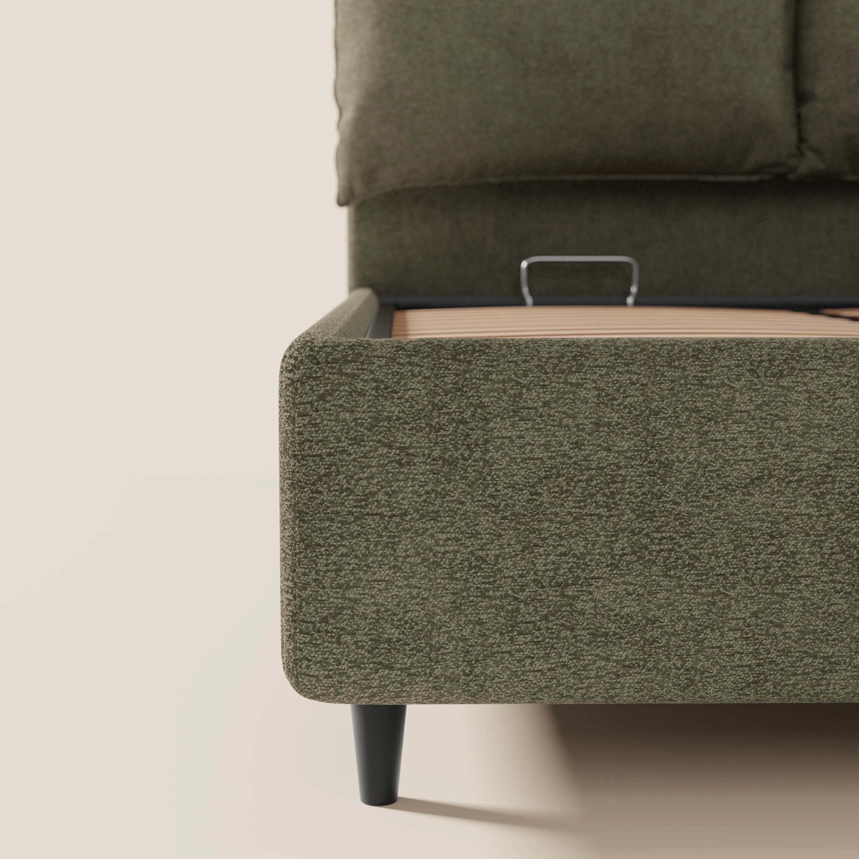 Primo piano del letto imbottito ZAYRON di Letti.store in tessuto verde oliva certificato Oeko-Tex®, caratterizzato da morbidi cuscini regolabili, un angolo arrotondato, gambe affusolate nere, base a doghe in legno con vano contenitore, il tutto su sfondo di pareti e pavimento beige chiaro.
