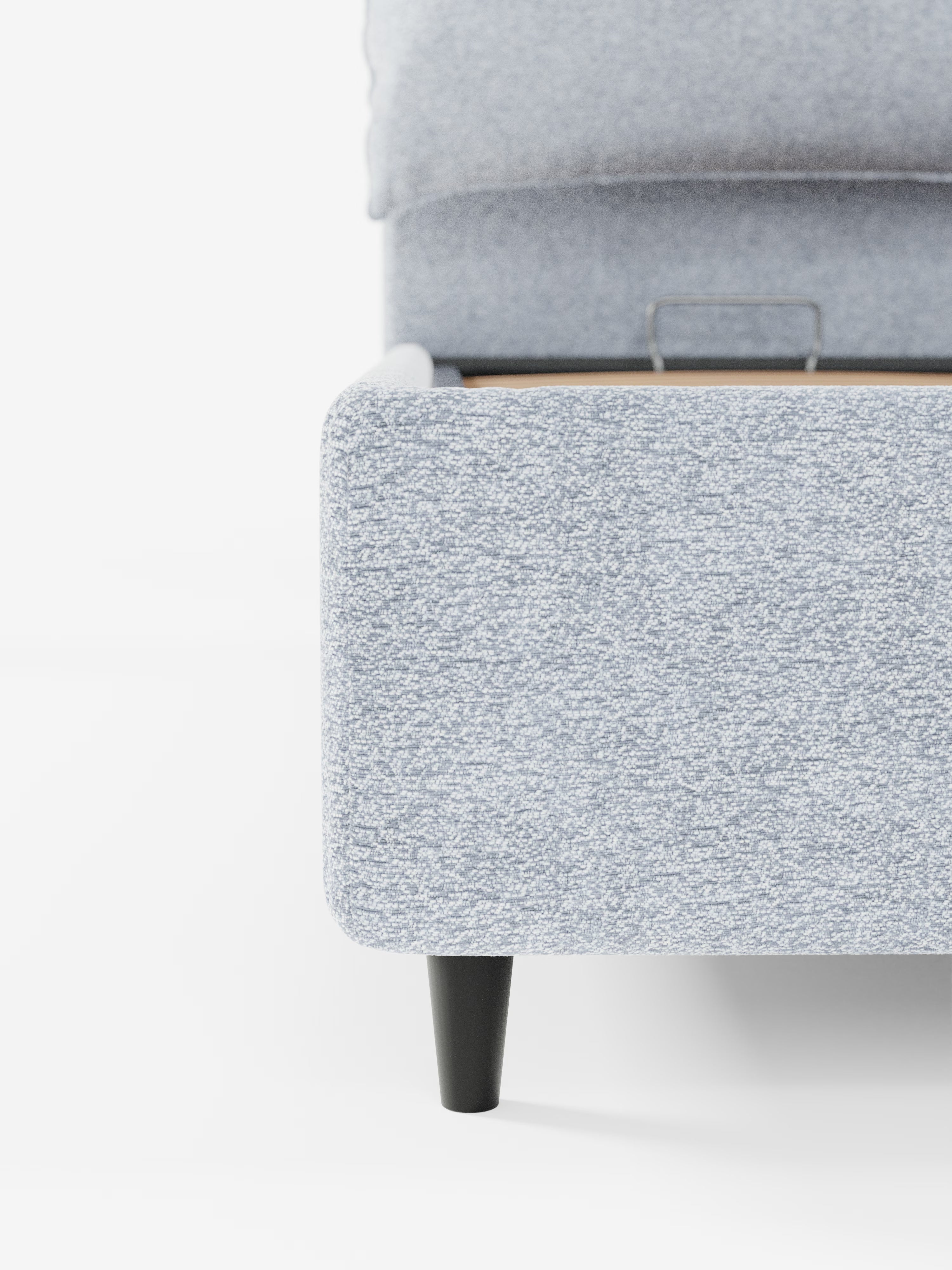 Primo piano di un letto imbottito ZAYRON grigio chiaro con vano contenitore e morbidi cuscini regolabili di Letti.store, caratterizzato da una gamba rotonda in legno scuro su sfondo bianco. La parte superiore del letto è aperta, mettendo in evidenza l’ampio vano contenitore con maniglia in metallo integrata all’interno. Il design complessivo è moderno e minimalista, realizzato in tessuto certificato Oeko-Tex®.