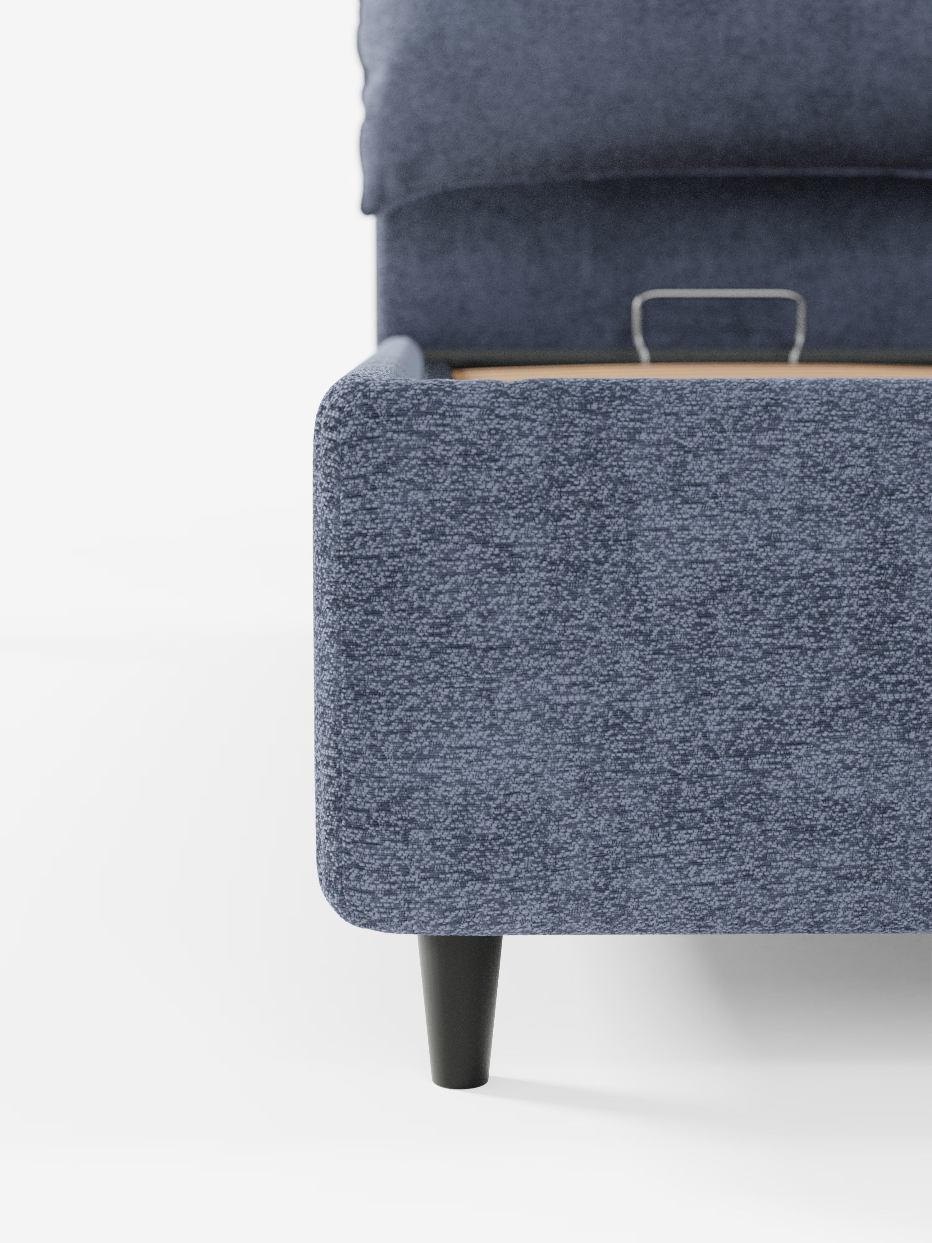 Primo piano di un angolo del letto imbottito ZAYRON con contenitore e morbidi cuscini regolabili di Letti.store, caratterizzato da un rivestimento in tessuto blu certificato Oeko-Tex® e da un'unica gamba in legno nero. È visibile la trama del tessuto dal design moderno, insieme a una parte della testiera regolabile e della base del letto, il tutto su uno sfondo bianco a tinta unita per mettere in risalto il mobile.