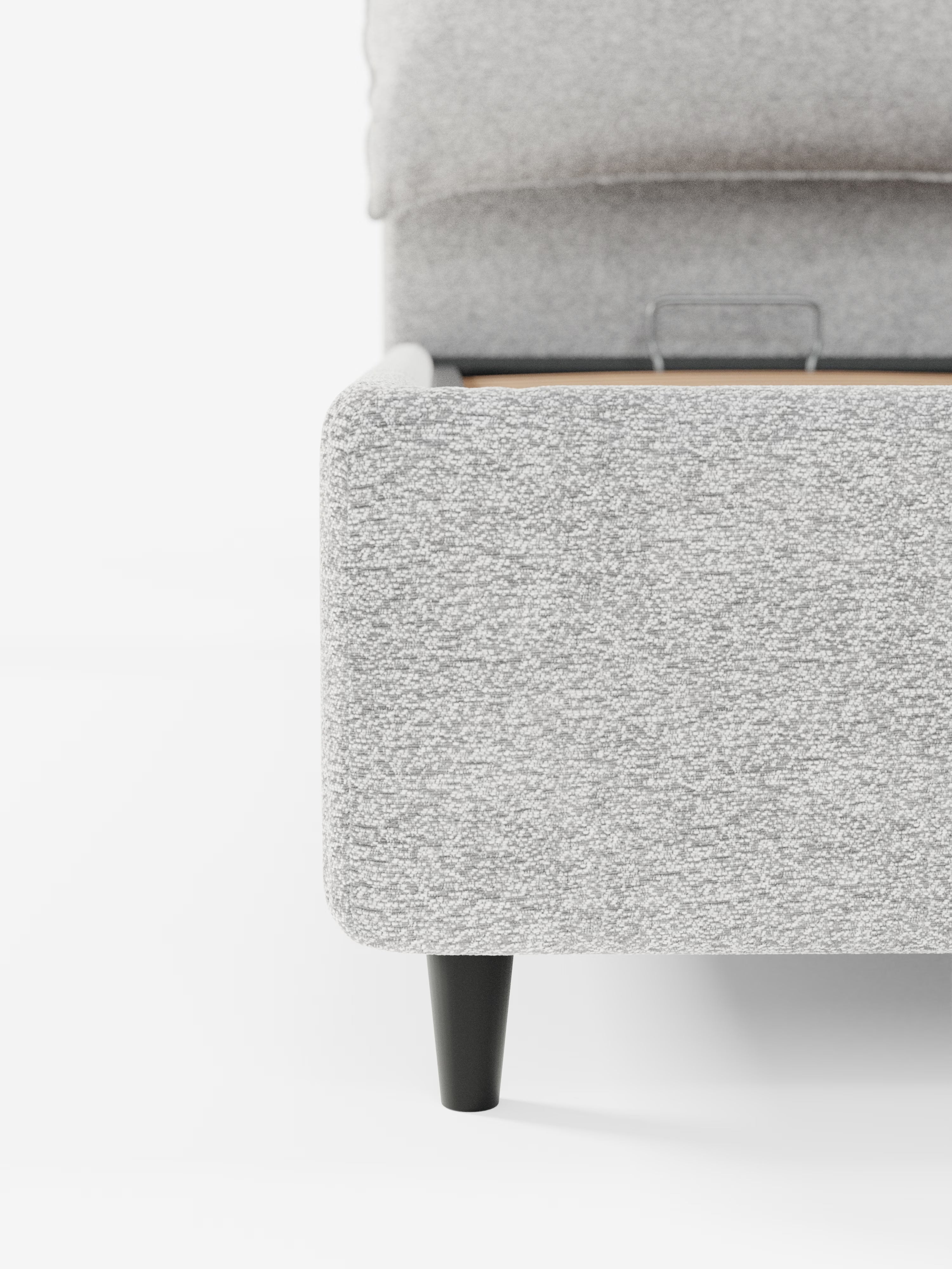 Primo piano dell'angolo del letto imbottito ZAYRON con contenitore e morbidi cuscini regolabili di Letti.store, caratterizzato da un moderno tessuto grigio chiaro certificato Oeko-Tex®, una gamba affusolata nera e doghe in legno parzialmente visibili. Lo sfondo bianco minimalista ne esalta il design pulito e contemporaneo.
