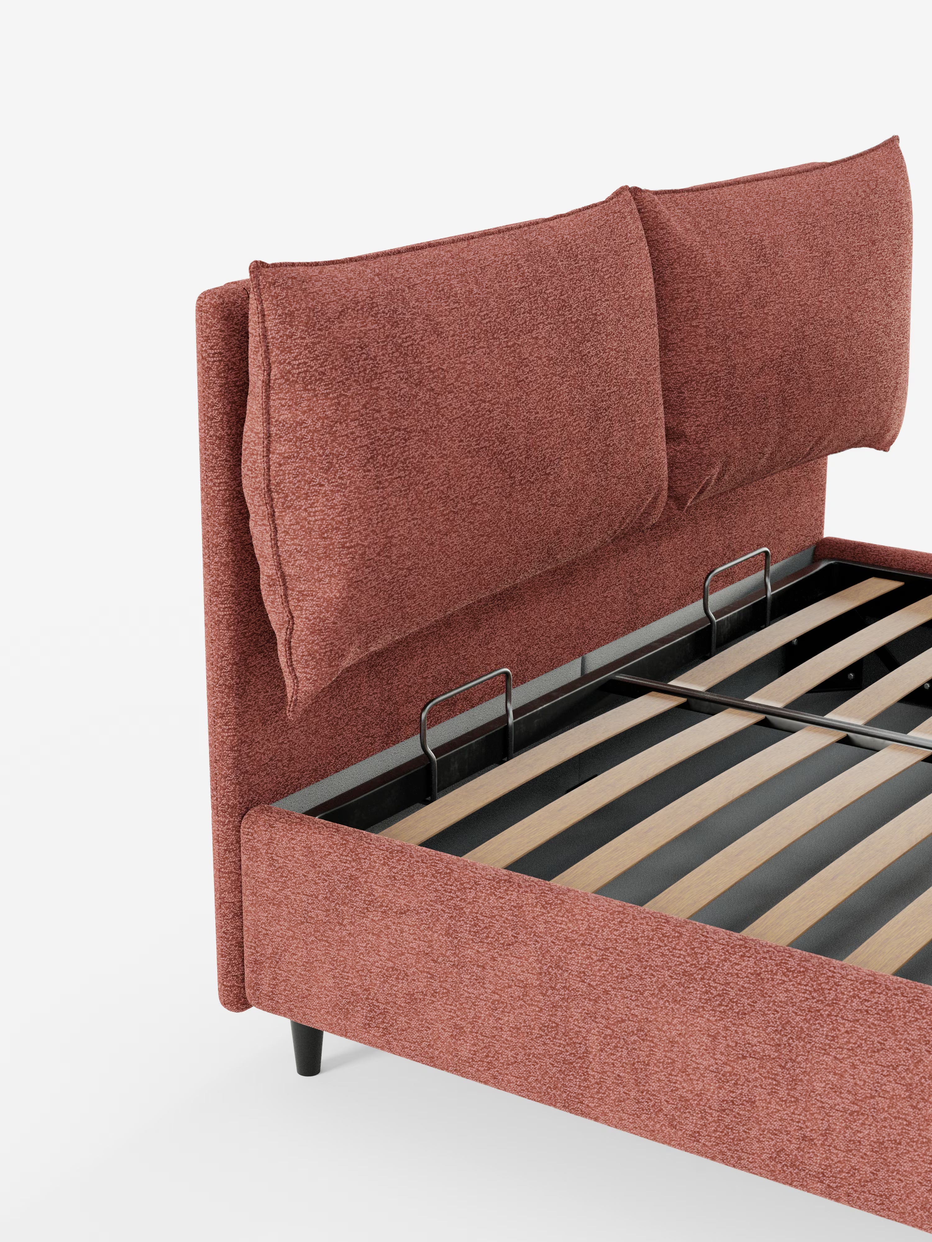 Una vista parziale del letto imbottito ZAYRON con contenitore e morbidi cuscini regolabili di Letti.Il modello, presentato nel negozio, presenta un rivestimento in tessuto color ruggine certificato Oeko-Tex®, una testiera imbottita con due grandi cuscini regolabili, doghe in legno a vista e montanti in metallo nero, il tutto su uno sfondo bianco a tinta unita.