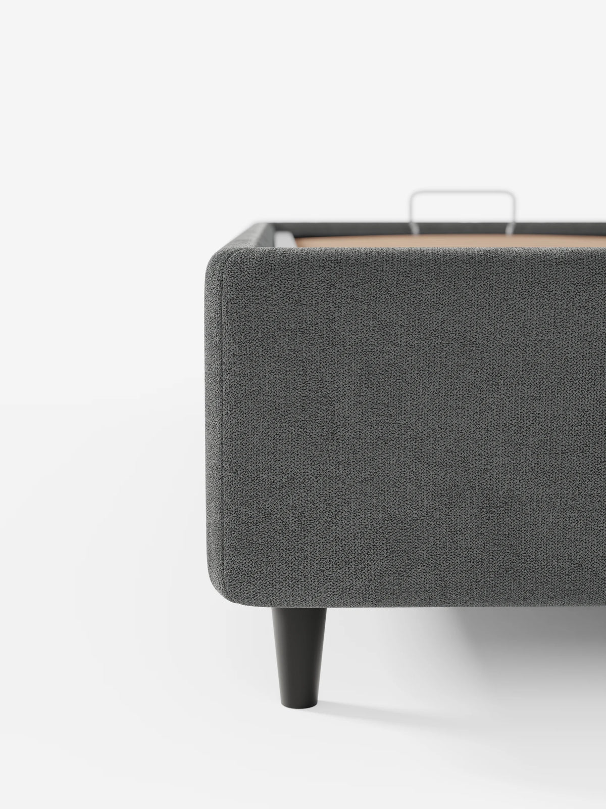 Un primo piano di un letto sommier ERON imbottito di colore grigio, dotato di vano contenitore in tessuto impermeabile certificato Oeko-Tex, caratterizzato da una base in tessuto testurizzato su gambe corte e scure, con una sottile maniglia in metallo sulla piattaforma.