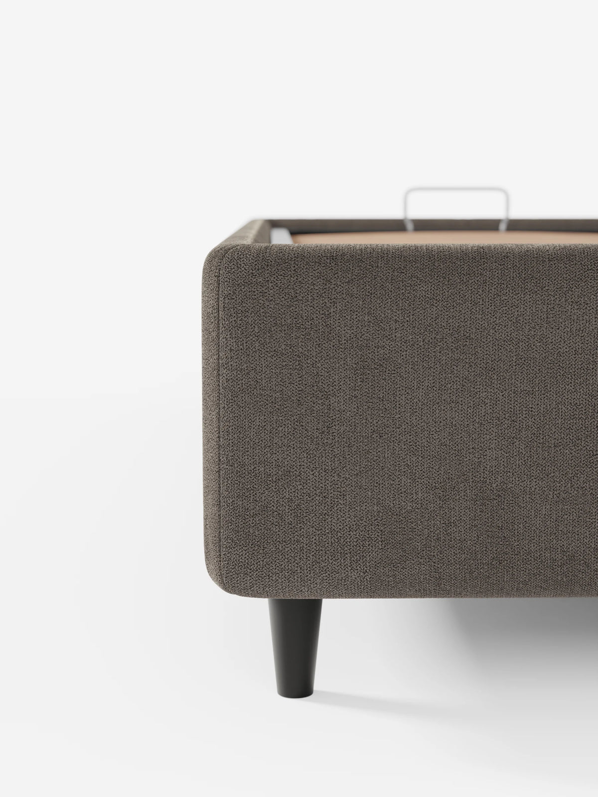 Un primo piano di un letto sommier ERON grigio scuro, imbottito e dotato di vano contenitore in tessuto impermeabile certificato Oeko-Tex, con bordi arrotondati, gambe affusolate e una maniglia in metallo visibile sulla piattaforma piana all’interno del telaio.