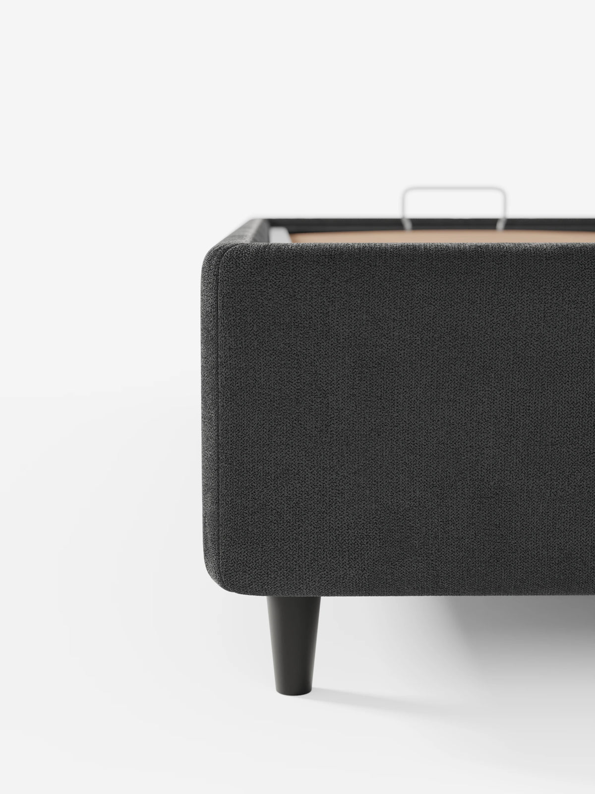 Un primo piano dell'angolo di un letto sommier imbottito ERON grigio scuro con vano contenitore in tessuto impermeabile certificato Oeko-Tex, che mostra la trama del tessuto, una gamba nera corta e affusolata e una maniglia in metallo sulla base in legno.