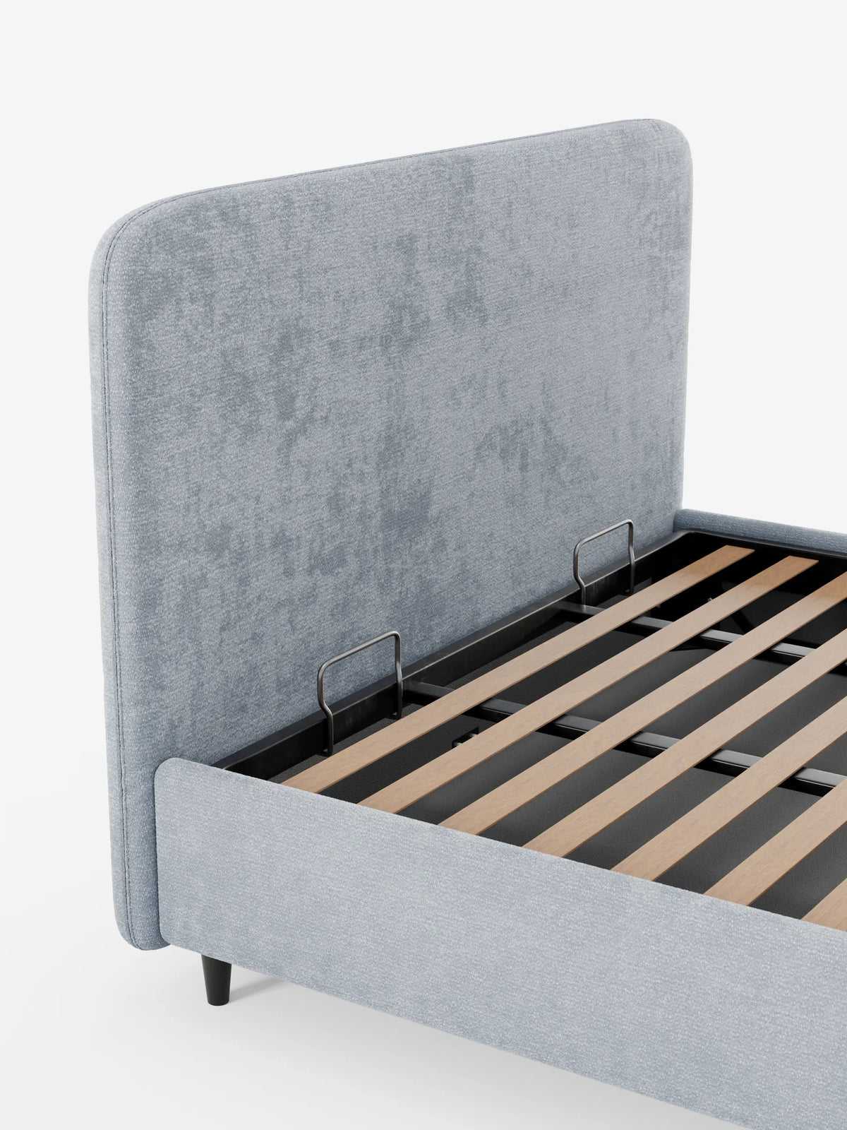 Un letto KAELOS minimal imbottito di colore grigio chiaro, con una testiera alta, arrotondata e in tessuto testurizzato, ripreso da un’angolazione dall’alto che ne rivela la base a doghe in legno e le due barre metalliche di fissaggio del materasso su uno sfondo bianco a tinta unita.