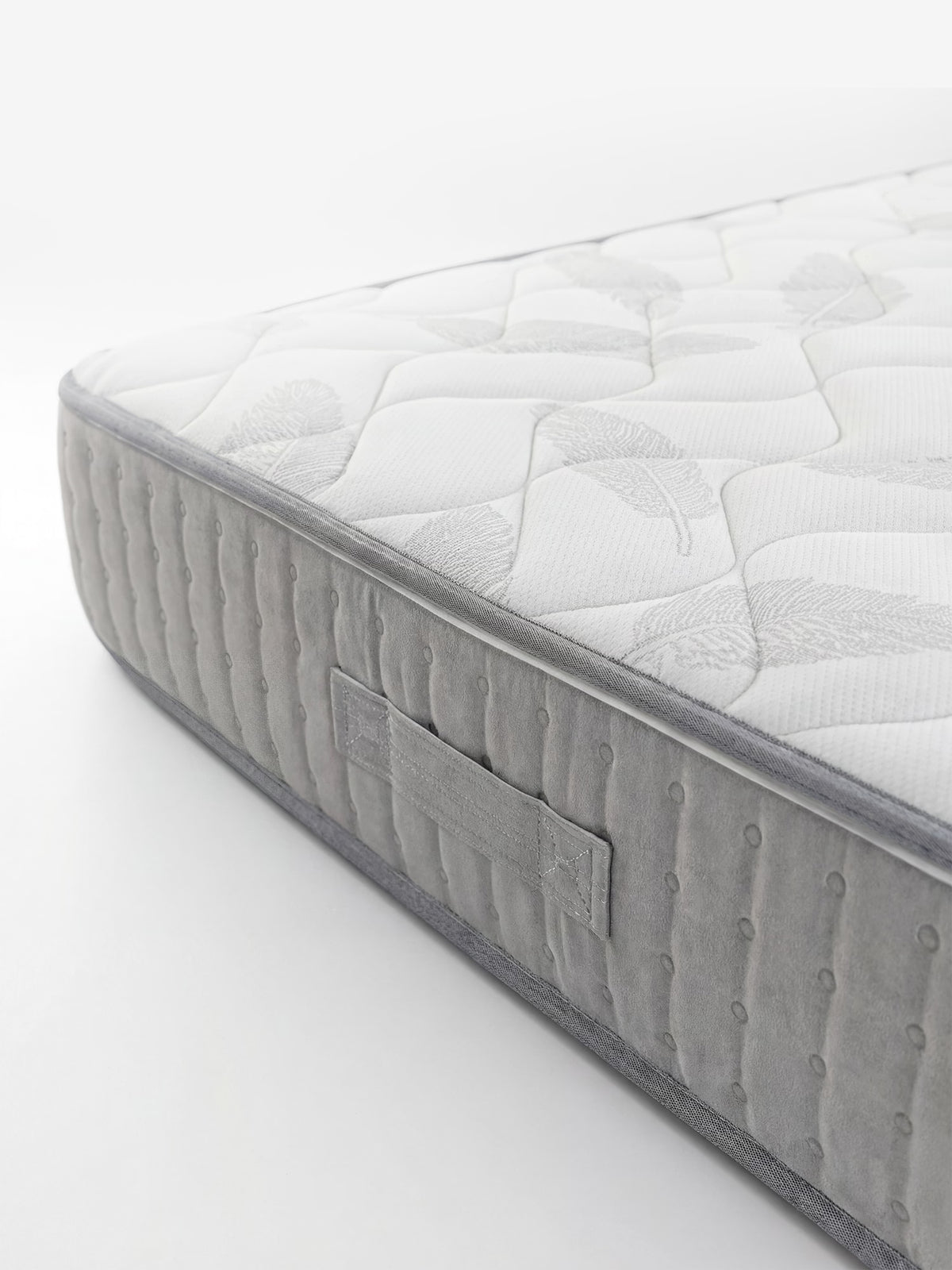 Primo piano del modello AERION – Materasso ortopedico in Memory & Aquapur per un riposo leggero come l’aria di Divani.Store, caratterizzato da una superficie superiore bianca trapuntata con motivo a piume e dai bordi in velluto grigio. Sul lato è cucito un manico in tessuto e l’angolo del materasso è visibile su uno sfondo bianco a tinta unita.
