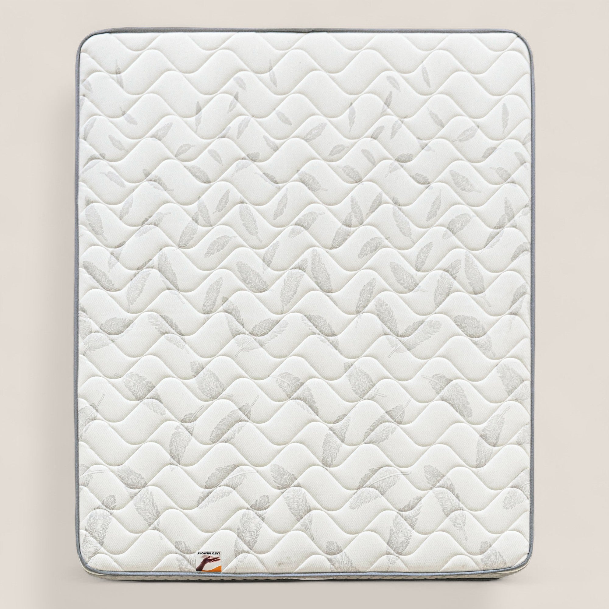 Il materasso ortopedico AERION Memory & Aquapur di Divani.Store è caratterizzato da una superficie trapuntata bianca con motivi ondulati e a foglia, bordature grigio chiaro e una piccola etichetta con logo con accenti rossi. Lo sfondo è in tinta unita beige.